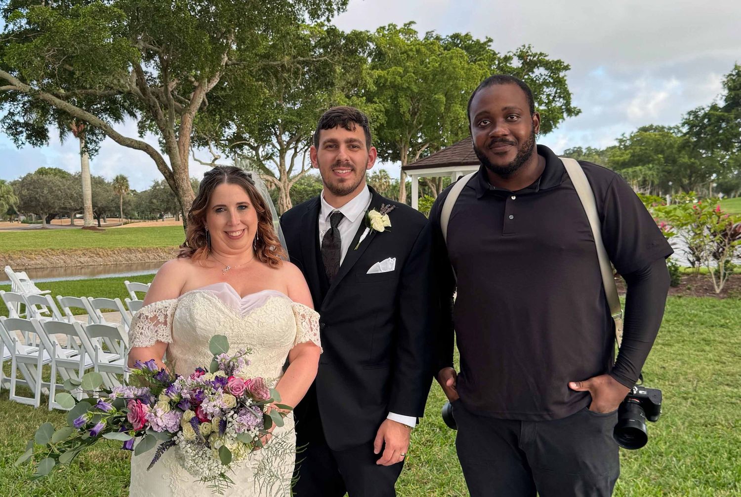 A wedding couple and photographer stand together on a grassy lawn with white chairs and trees in the background.