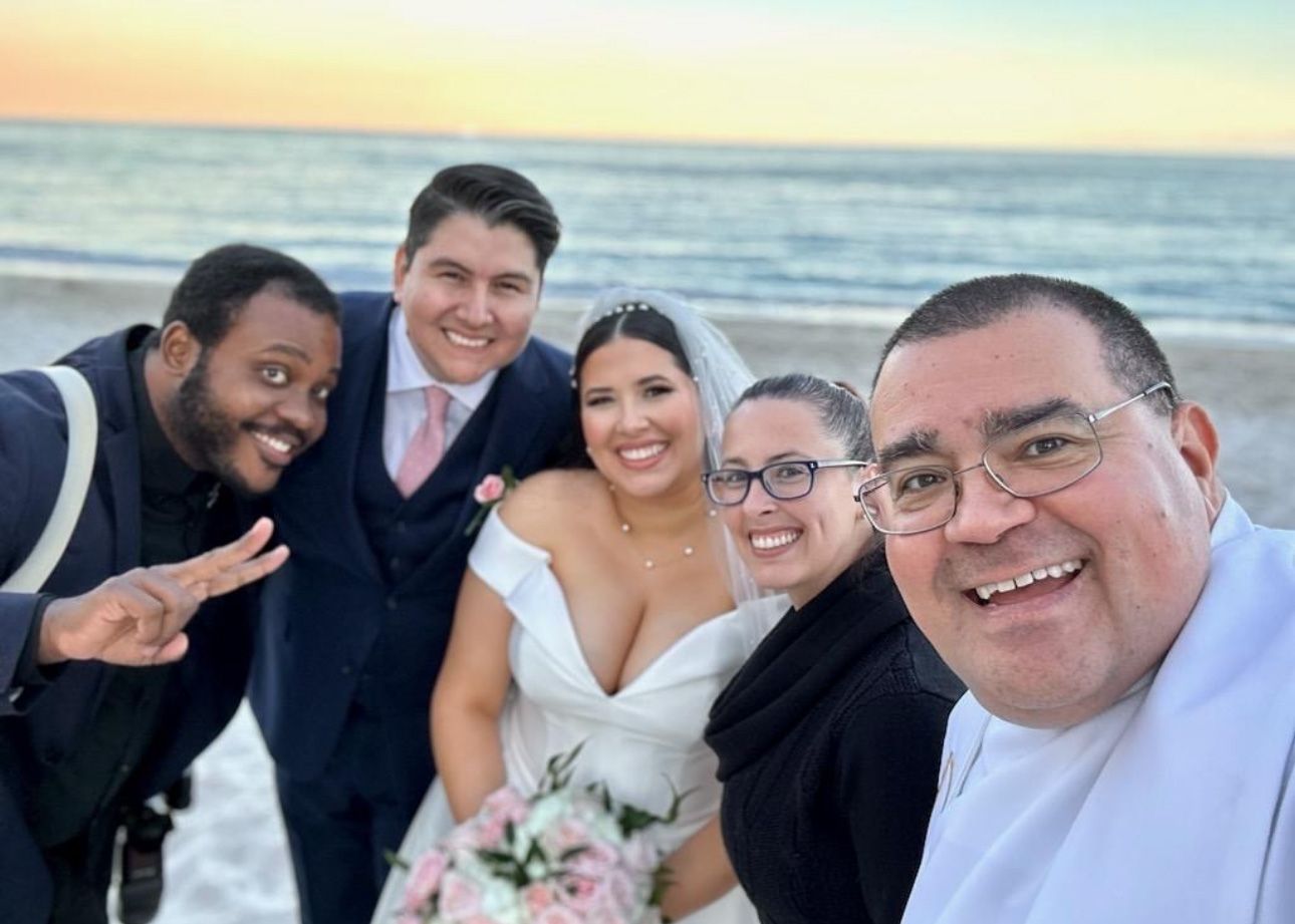 A group poses for a casual beach wedding selfie at sunset with the ocean visible in the background.