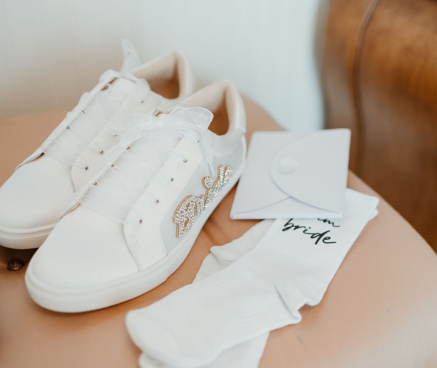 White leather bridal sneakers with delicate embellishments placed next to matching socks on a pink surface.