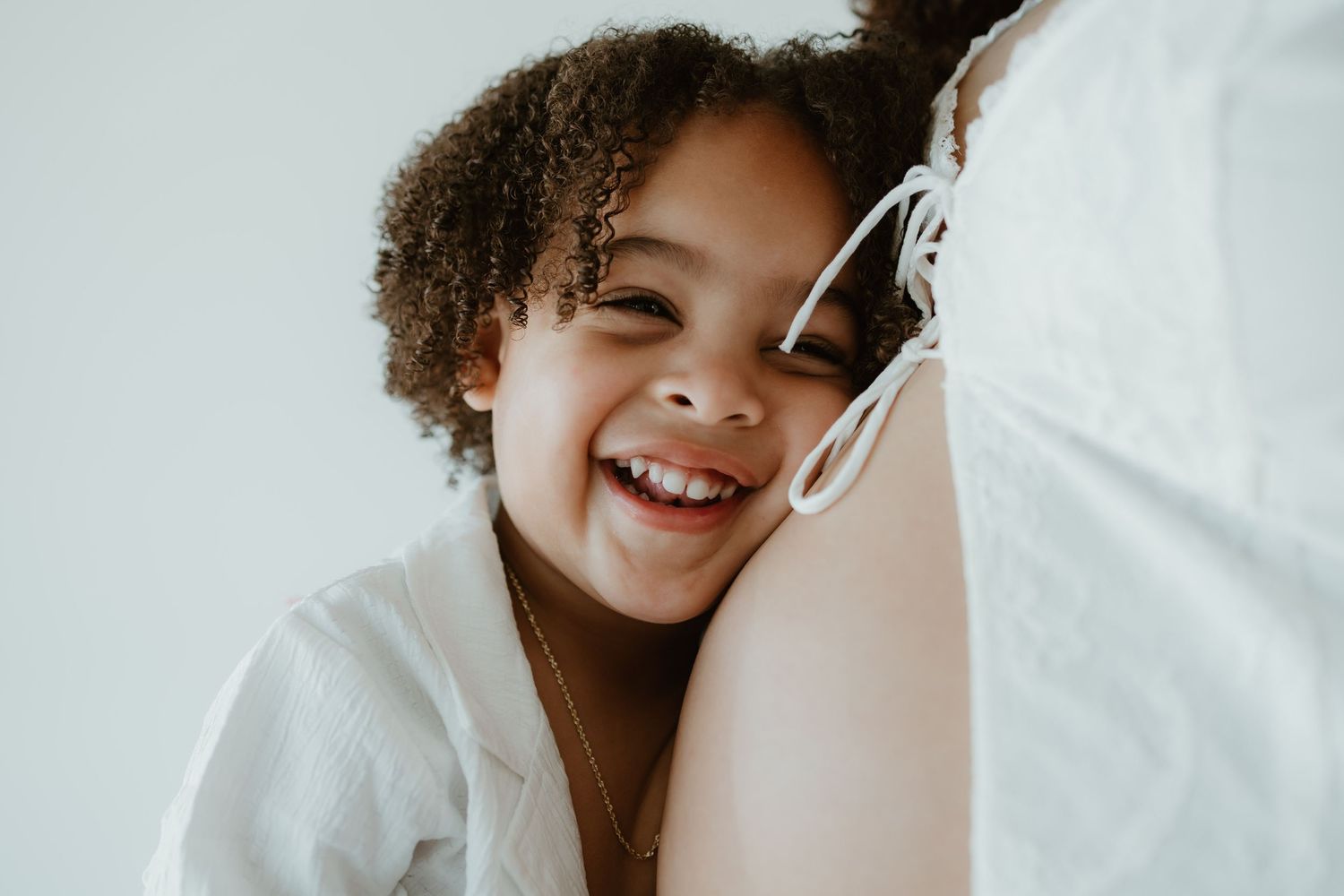 A joyful child with curly hair laughs while leaning against someone wearing white.