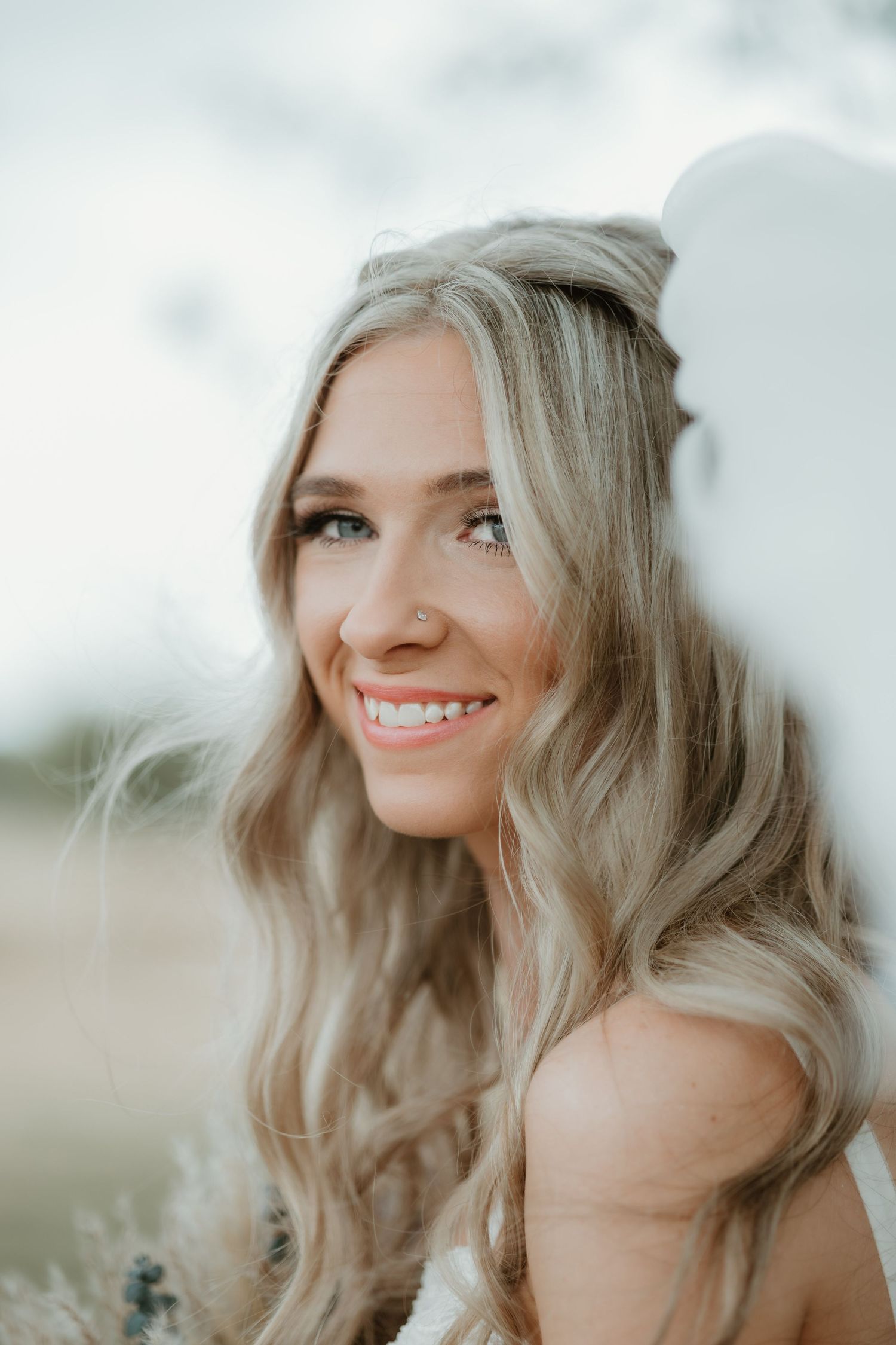 Close up of someone smiling warmly outdoors with wavy blonde hair and natural makeup in soft lighting.