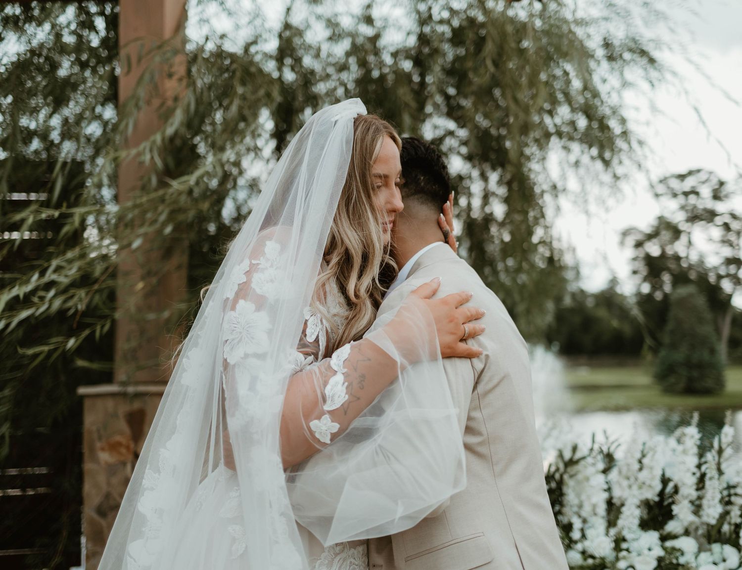 Two people share a romantic embrace in an outdoor wedding setting with a flowing white veil against willow trees and greenery.
