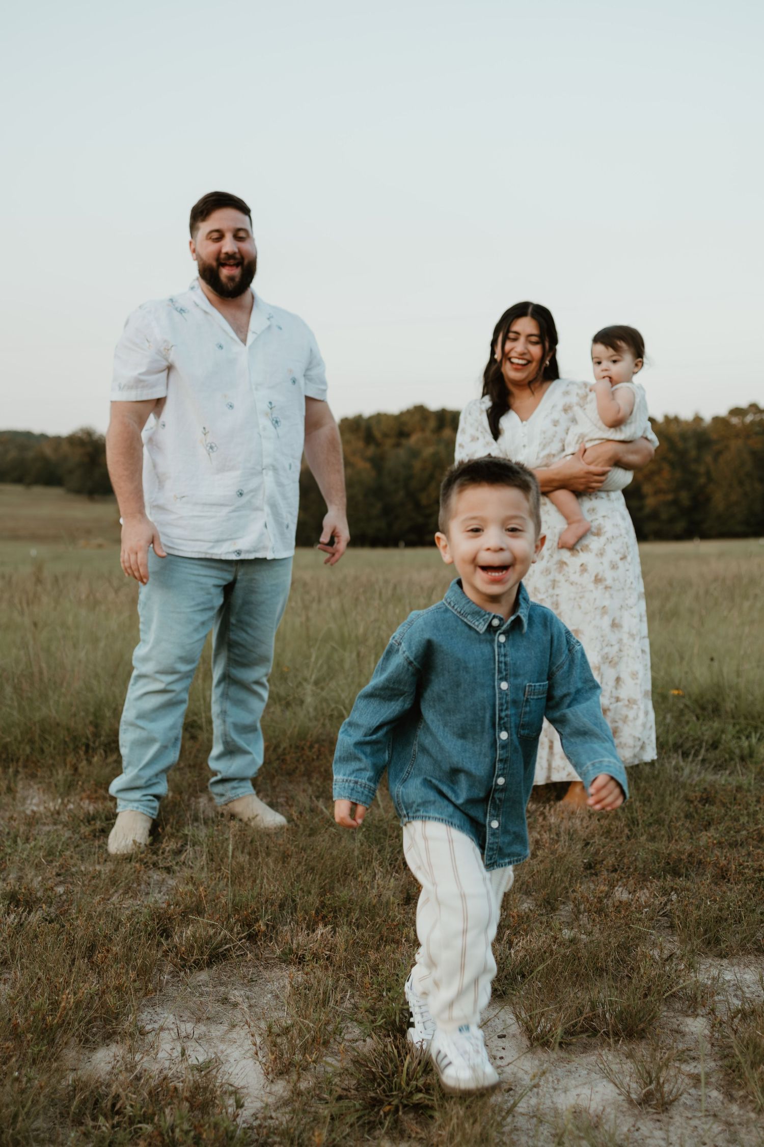 Family portrait in an outdoor field at sunset showing parents and two young children in casual attire.