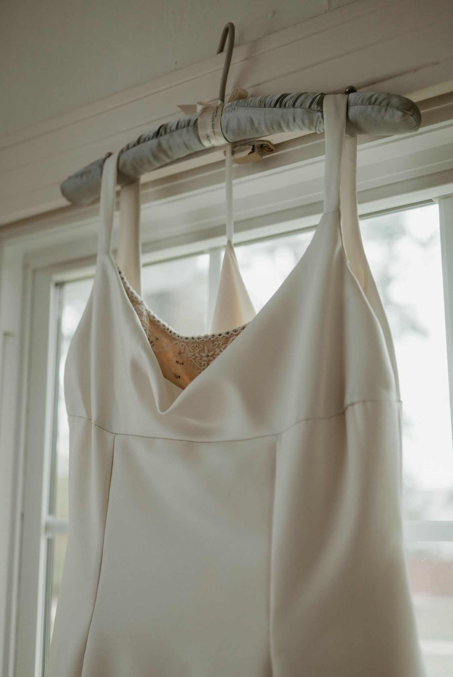 An elegant white wedding dress with a plunging neckline and delicate lace trim hangs by a window in natural light.
