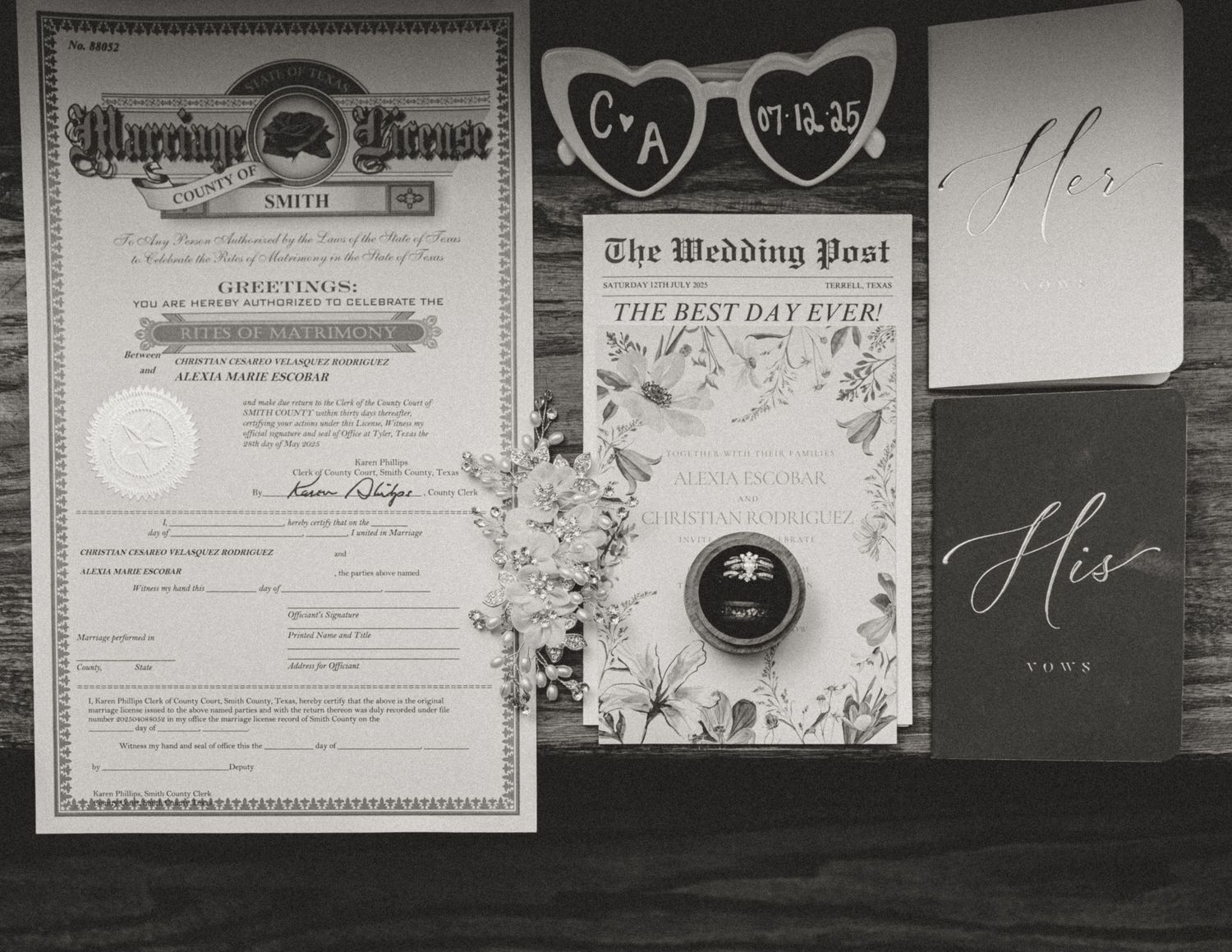 Wedding details flatlay with marriage license, invitation, rings, and floral elements on dark surface.