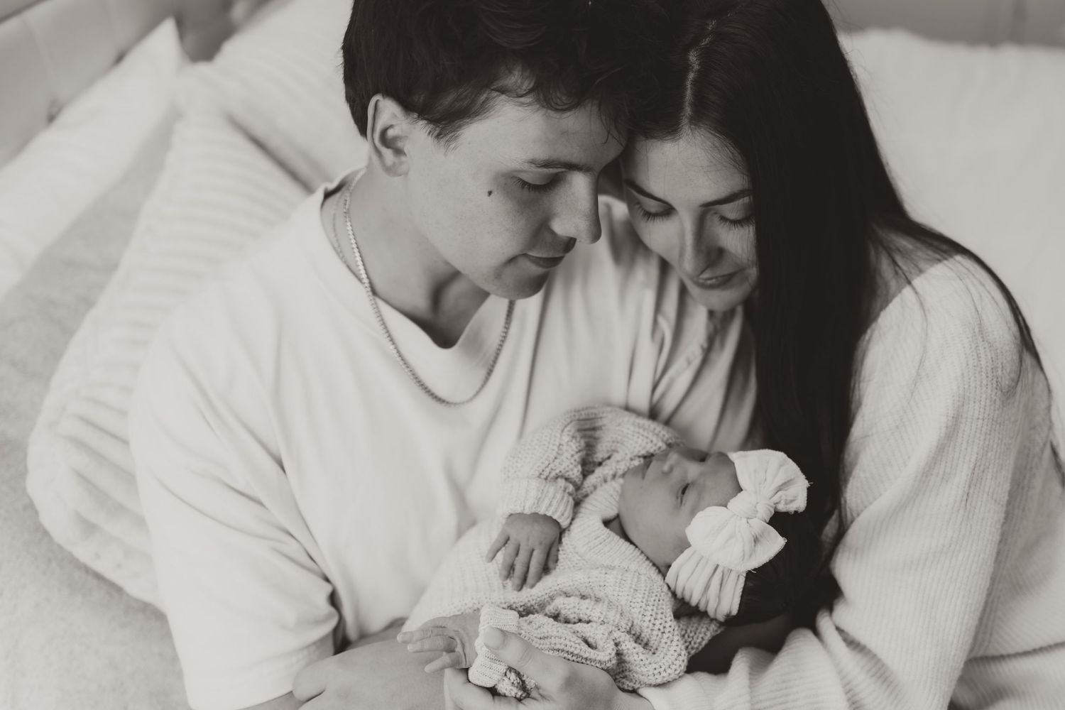 A black and white photo of new parents holding and gazing lovingly at their newborn baby wrapped in a blanket.