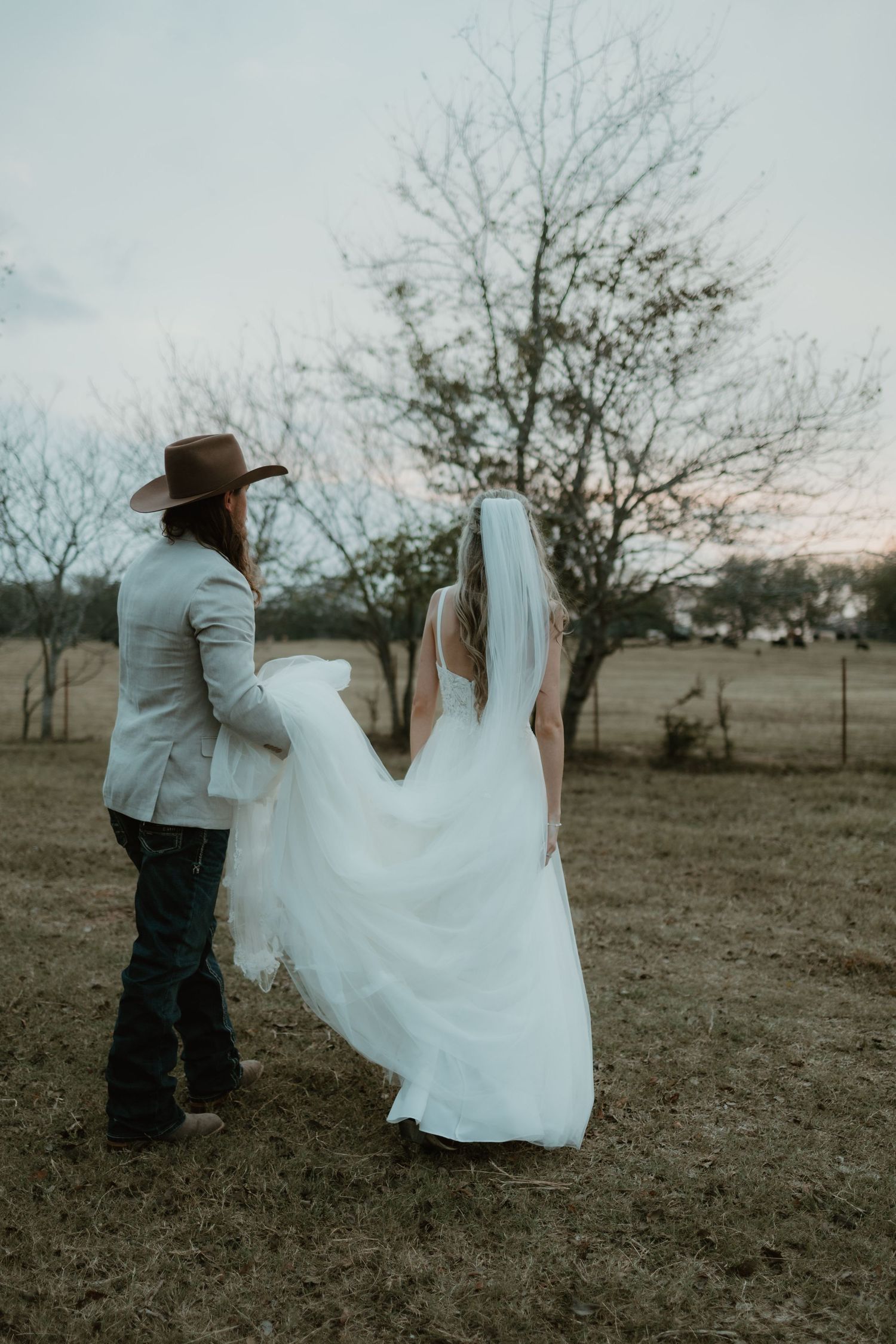 A rustic country wedding couple stands in a field at sunset with the bride's dress flowing in the breeze.
