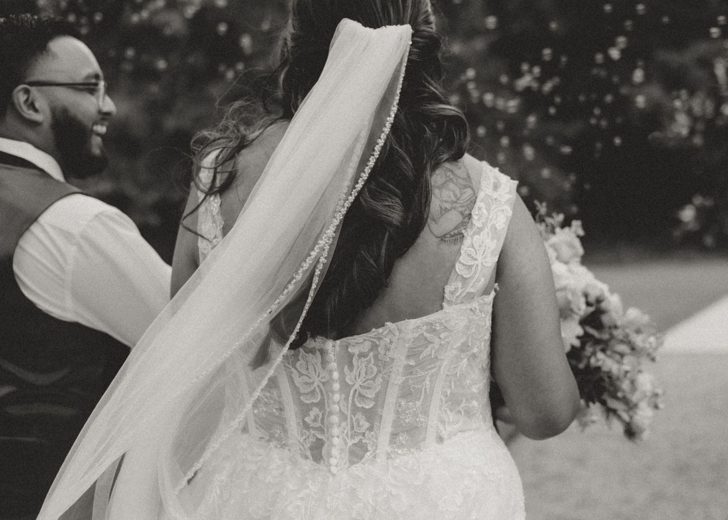 A close-up black and white photograph of a bride in a lace wedding dress with a flowing veil during an outdoor ceremony.