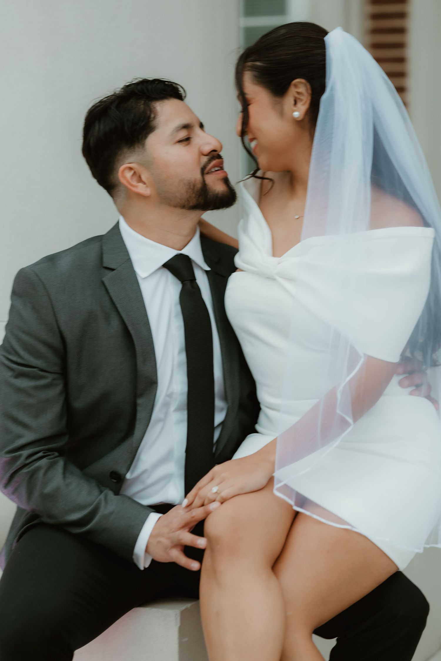 A romantic wedding portrait shows a bride in a white dress and veil sitting intimately with her groom in a gray suit.