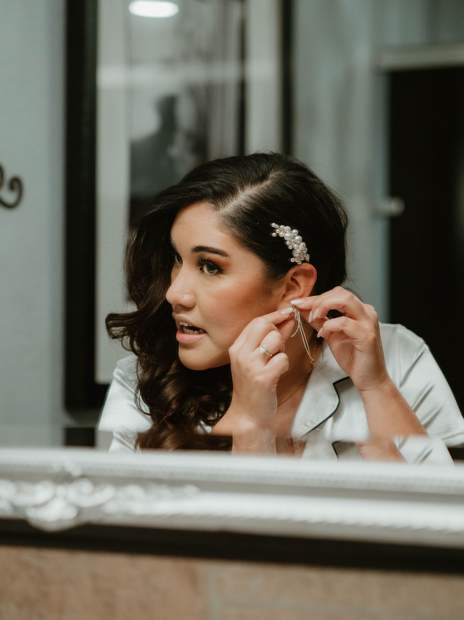 Bride putting on earrings in front of mirror while wearing elegant sparkly hairpiece and white dress.