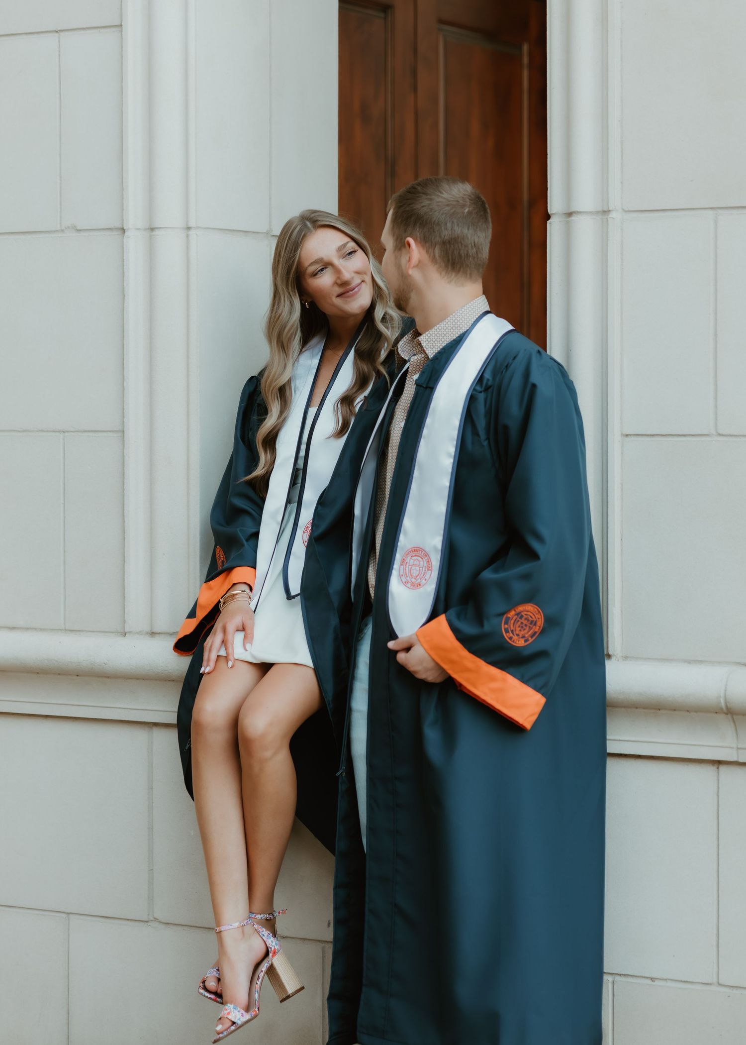 Two graduates in matching navy blue graduation gowns and caps share a candid moment against a white stone wall.