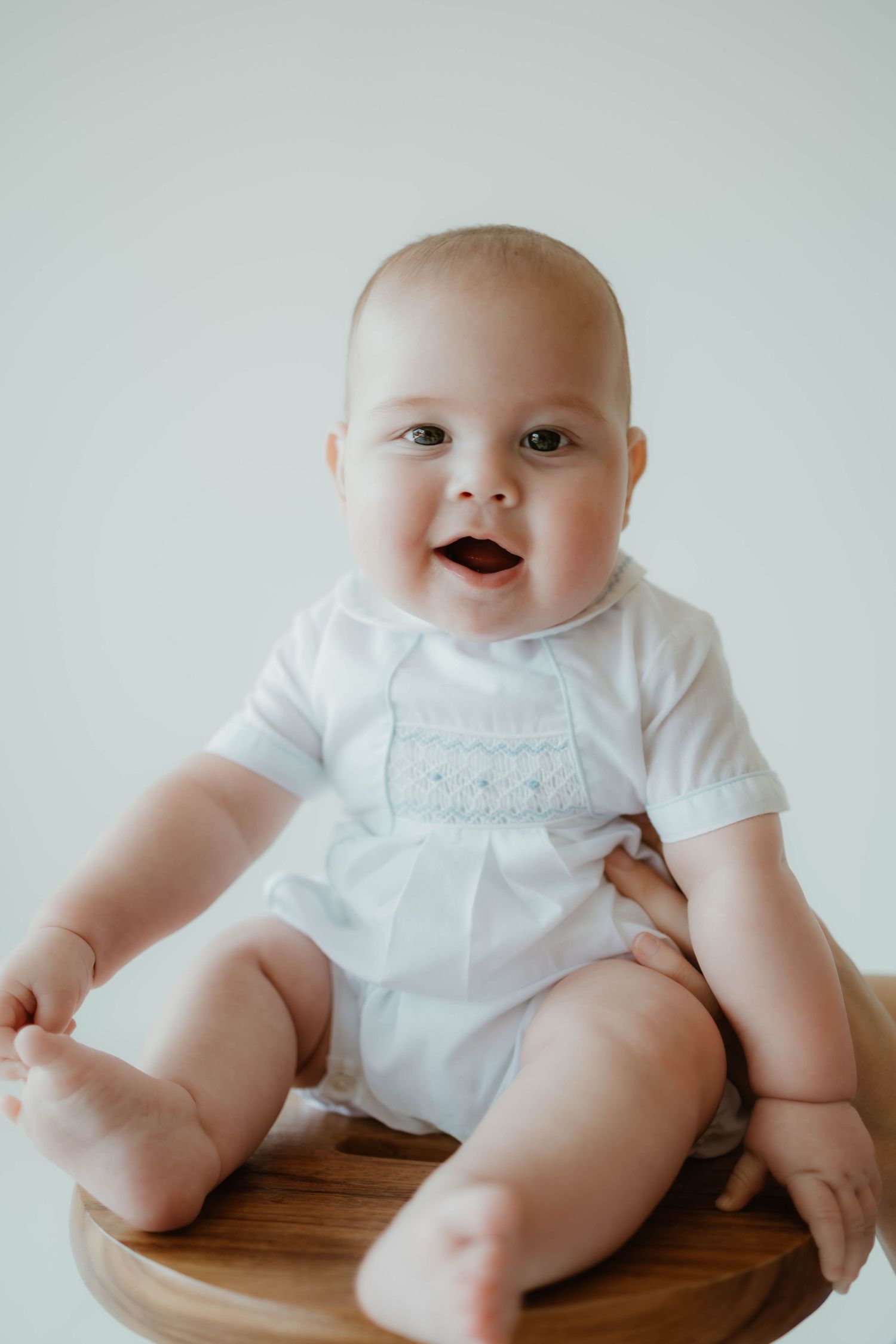 A happy baby wearing a white smocked outfit sits on a wooden surface against a light background.