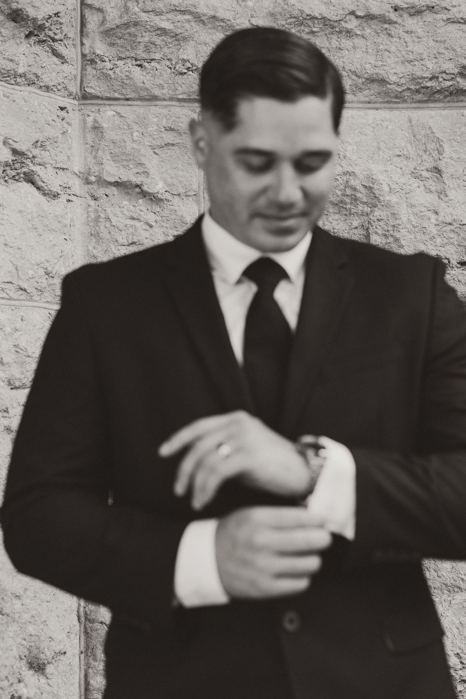 Black and white photo of a person in formal attire checking their watch while standing against a stone wall.