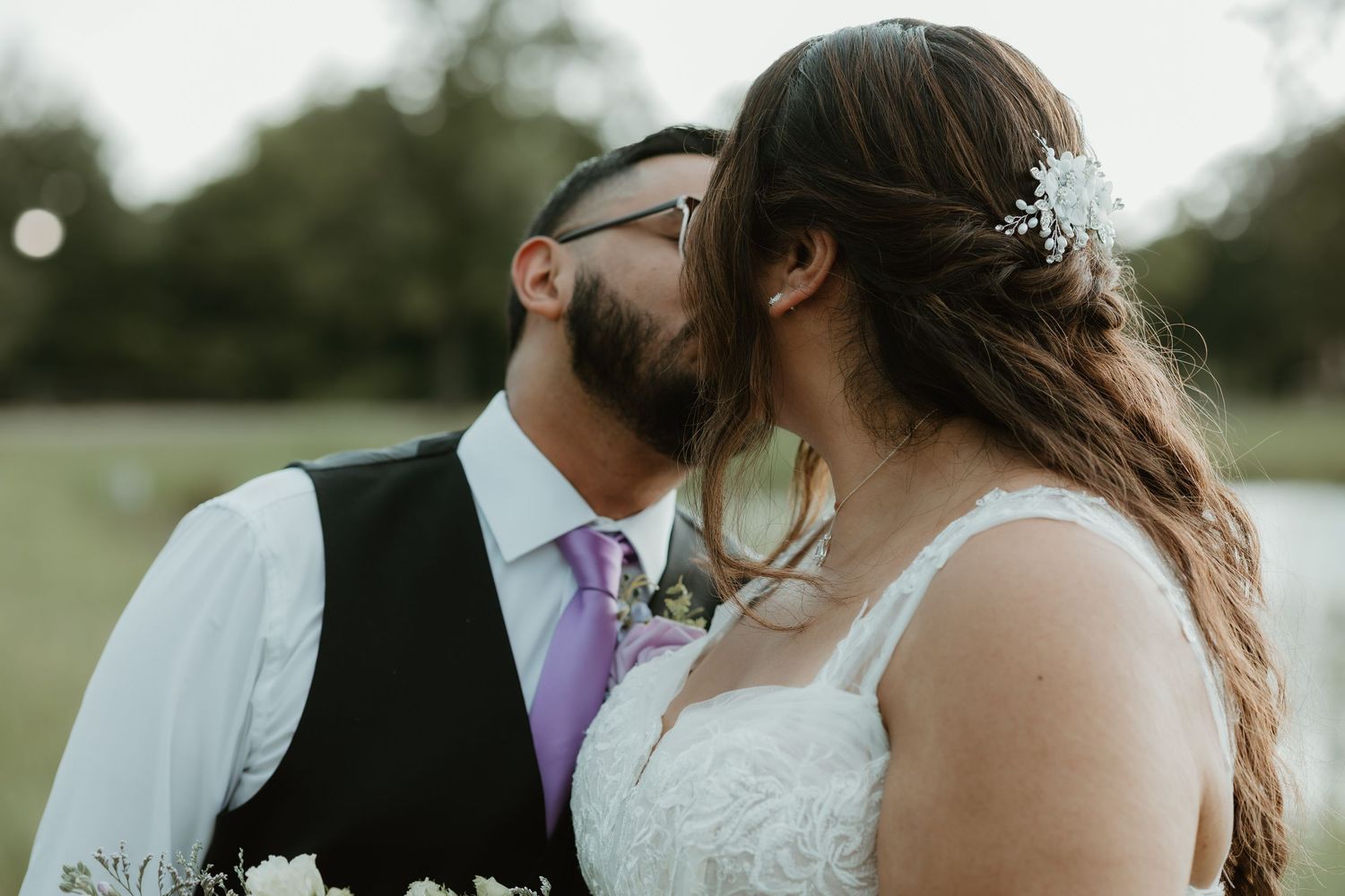 A couple shares a tender kiss during their outdoor wedding ceremony, with the groom in a vest and purple tie.