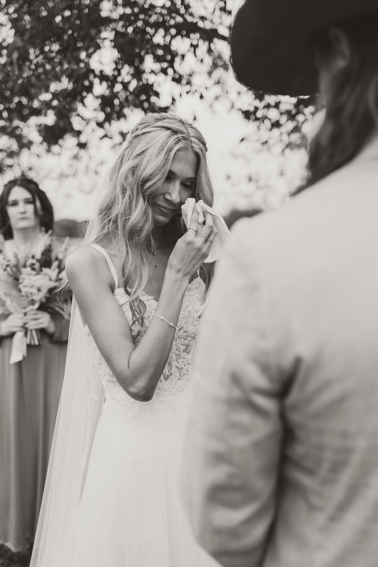 Emotional bride in white lace dress wipes tears during outdoor wedding ceremony while bridesmaid watches in background.