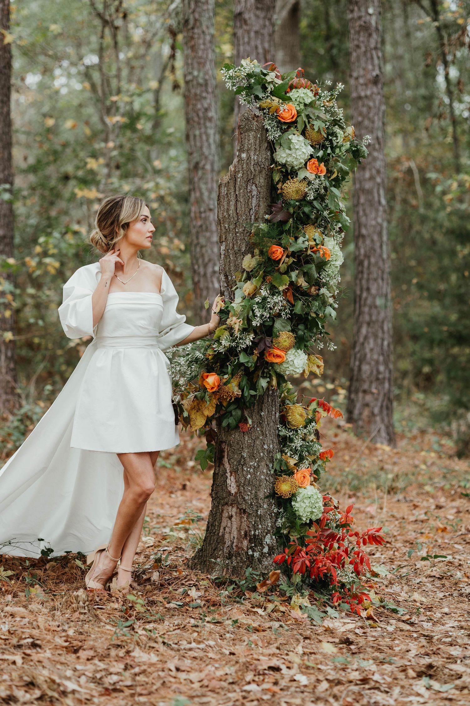 Floral arrangement with oranges and greenery decorates forest tree trunk beside figure in white bridal dress and bare feet.