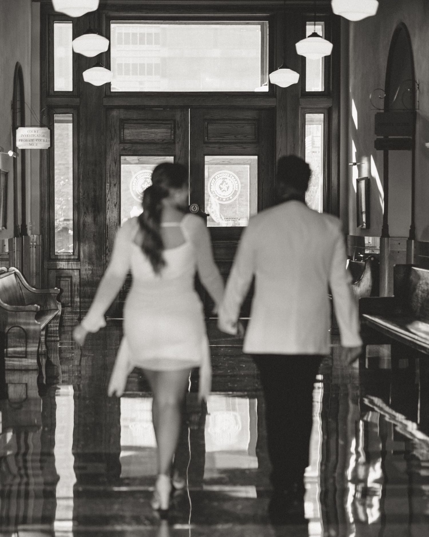 Black and white photo showing blurred silhouettes of people walking through a corridor with spherical light fixtures.