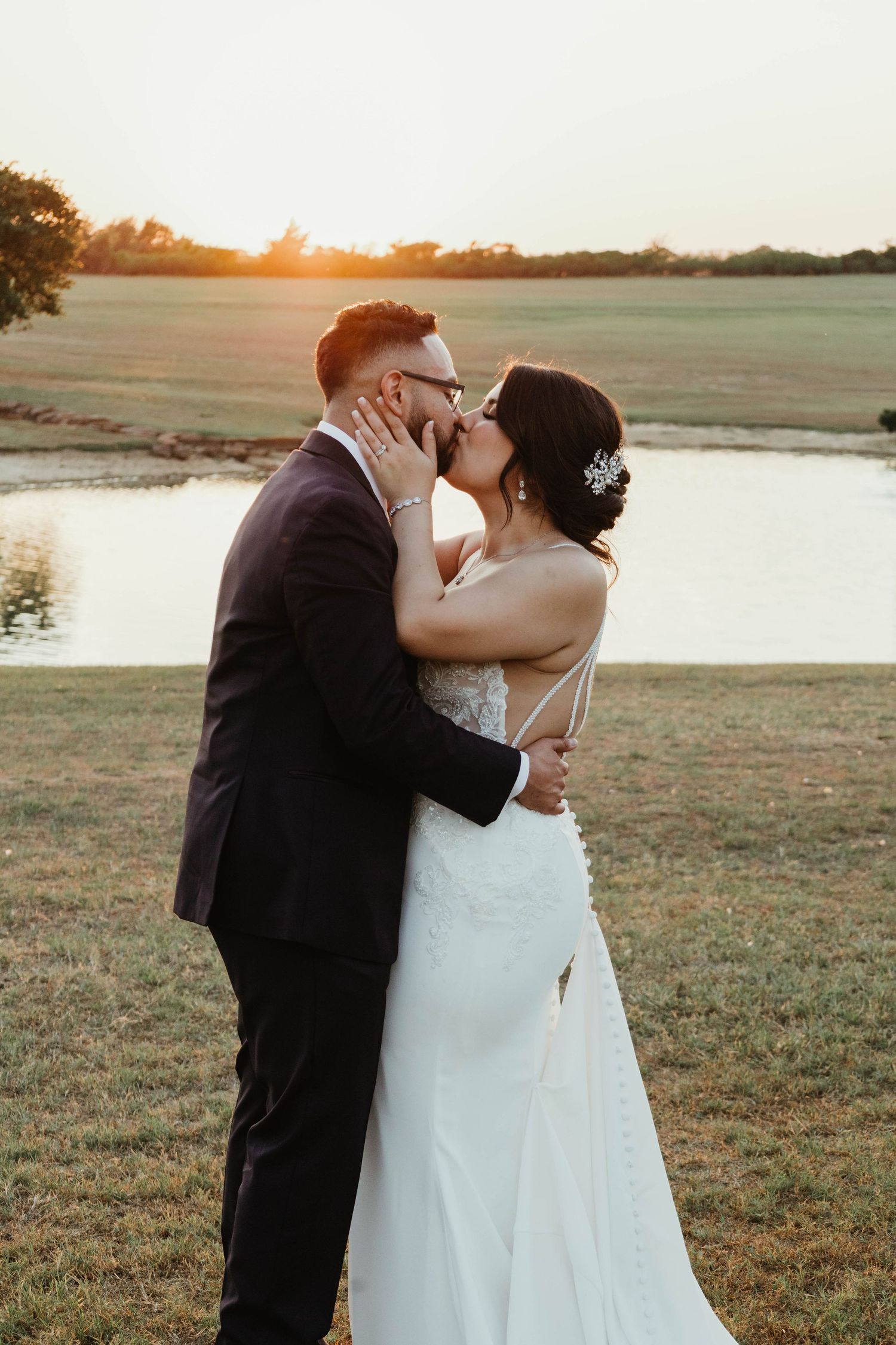 A romantic sunset wedding photograph of a couple sharing a kiss in an open field near a lake.