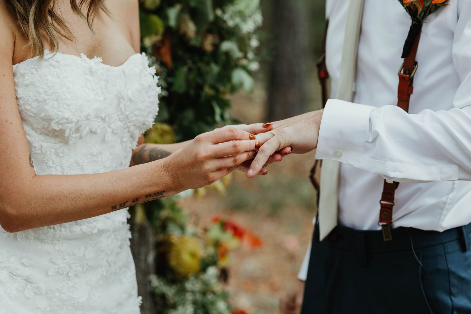 A close-up shot of hands exchanging wedding rings during a ceremony with white lace and floral decorations in background.