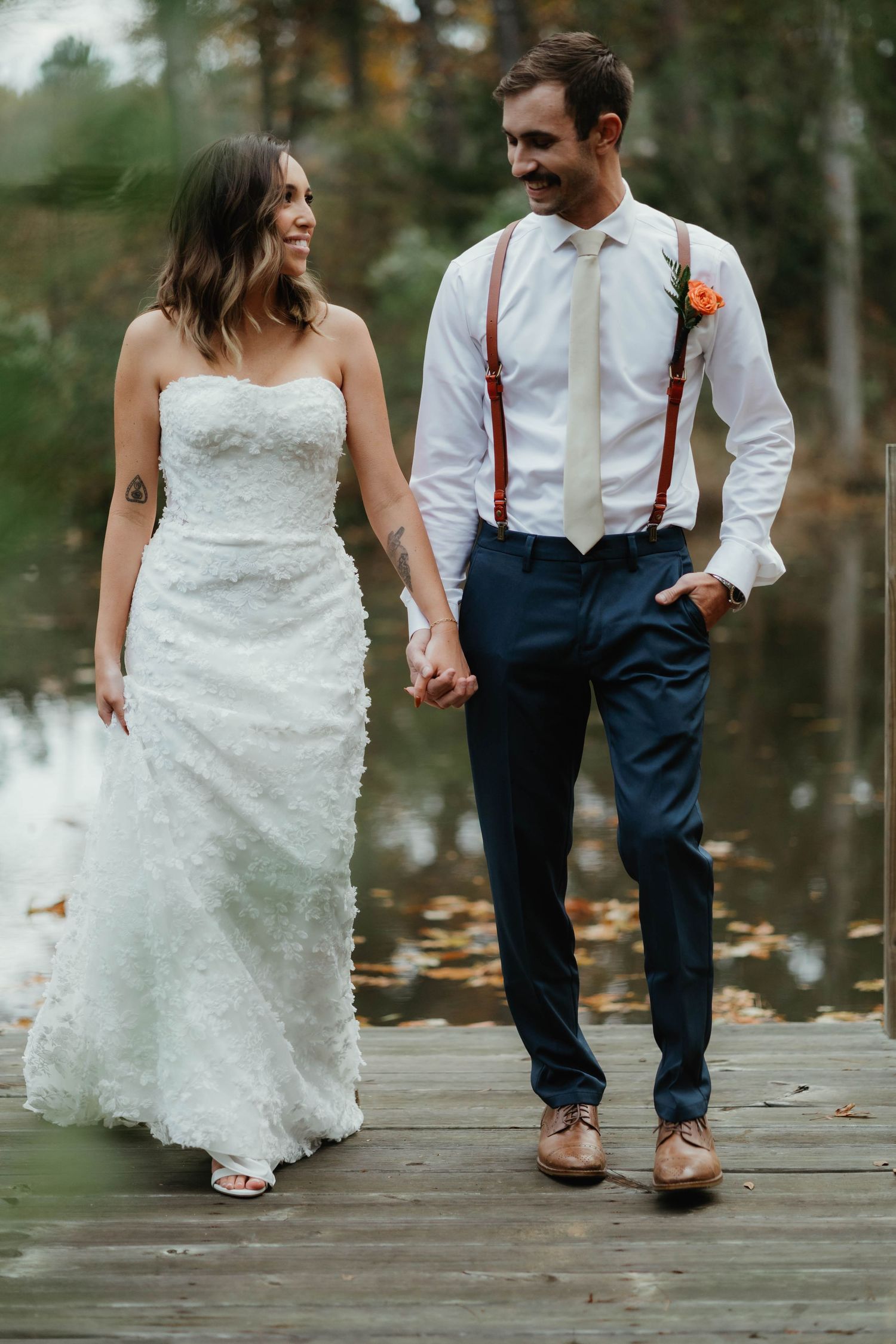 A couple walking hand in hand on a wooden dock, with the bride in a lace gown and groom wearing suspenders.