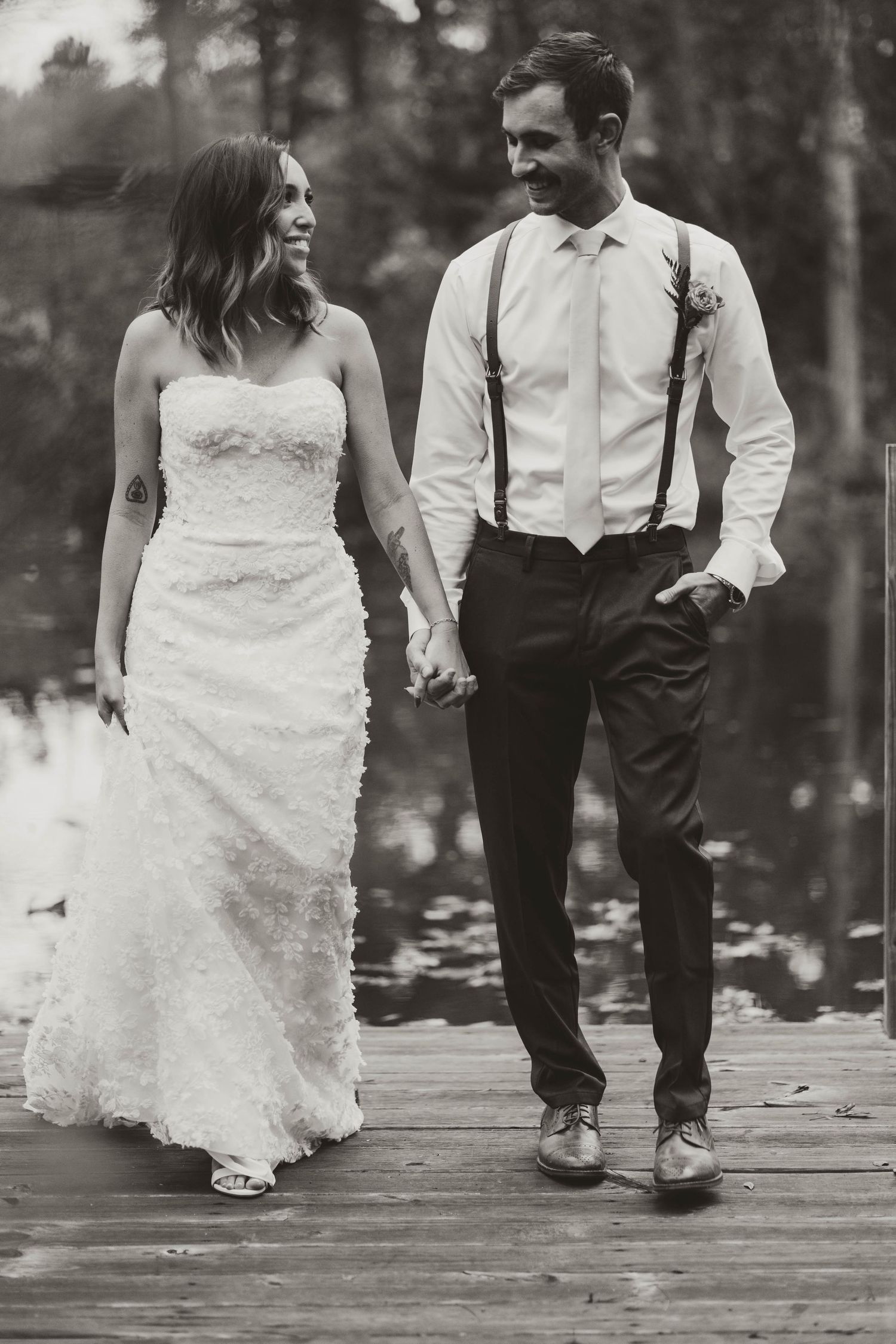 A black and white wedding photo of a couple holding hands while walking on a wooden deck in a forest setting.