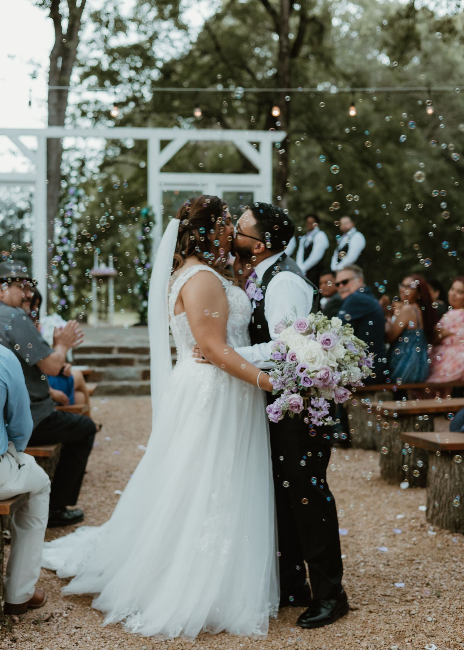 Newlyweds share their first kiss at an outdoor wedding ceremony while guests blow bubbles under string lights.