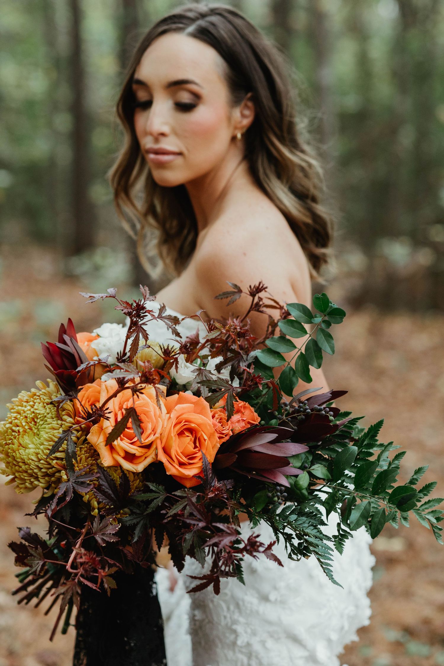 Romantic autumn wedding bouquet featuring orange roses, burgundy flowers and lush greenery in a moody forest setting.