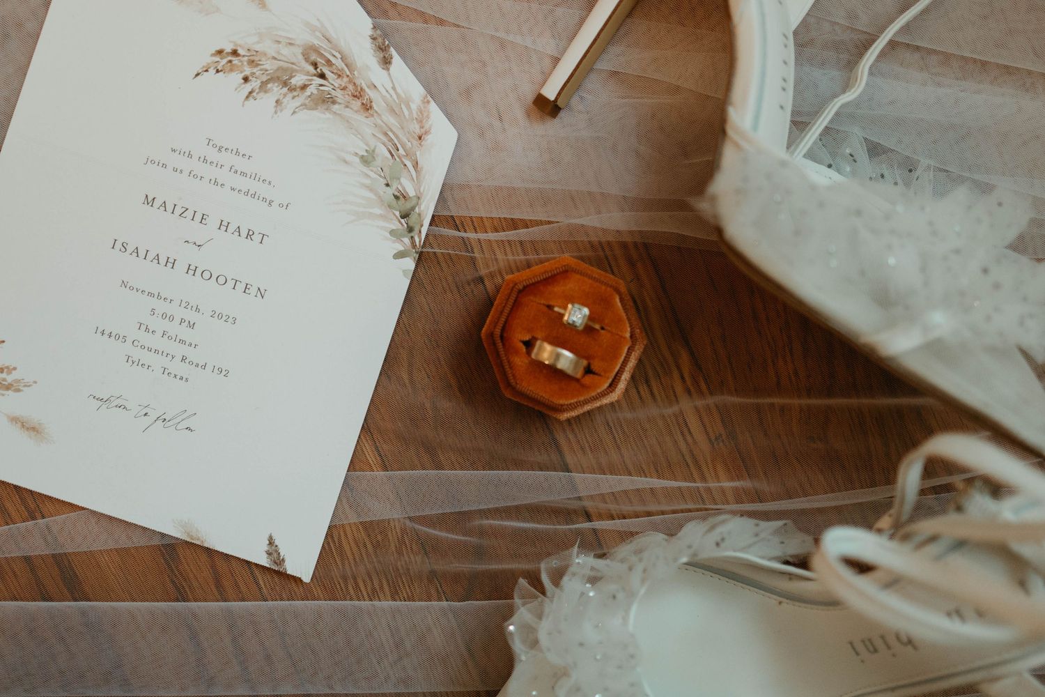 Wedding invitation and orange hexagonal ring box with jewelry on wooden surface beside veil and decor.
