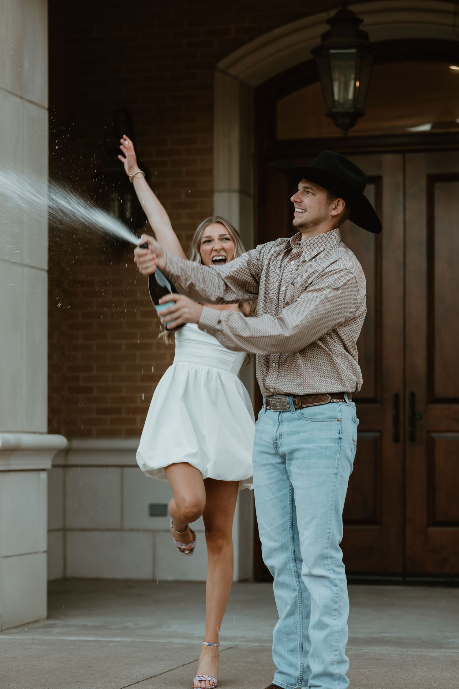 A celebratory couple pops champagne together outside a brick building with the woman in white and man in cowboy hat.