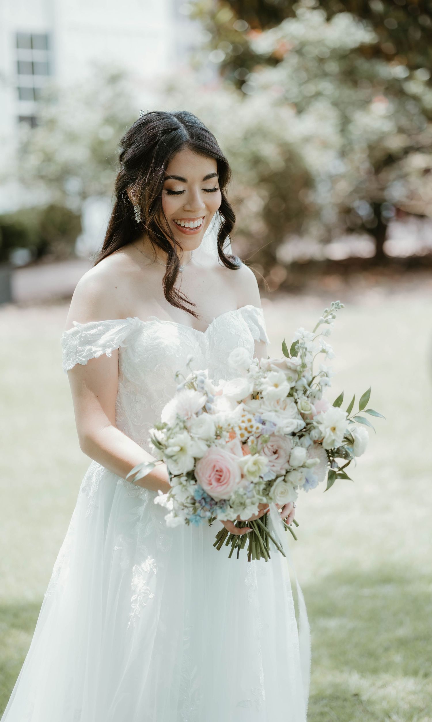 An elegant off-shoulder wedding dress with a delicate bouquet of pink roses and white flowers in an outdoor garden setting.