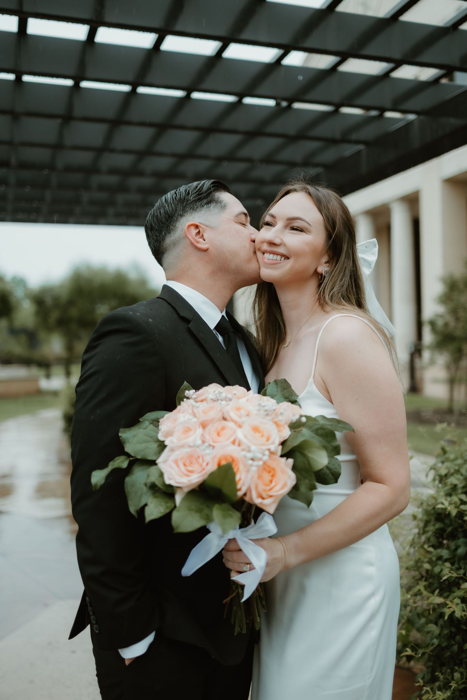 Newlywed couple shares intimate moment outdoors while bride holds peach rose bouquet with eucalyptus.