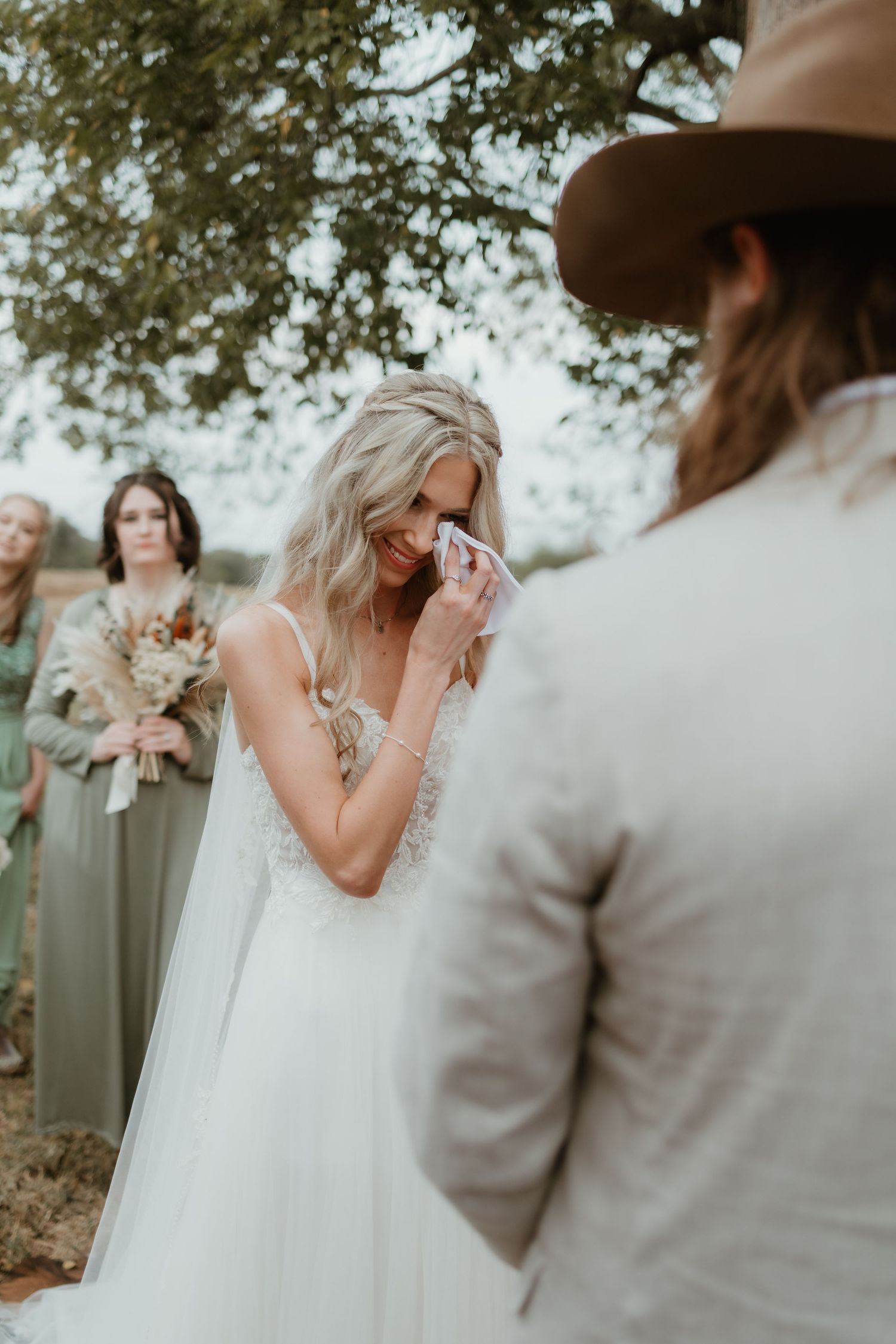 An emotional wedding ceremony moment shows a bride in white dress wiping tears as person in cowboy hat stands nearby.