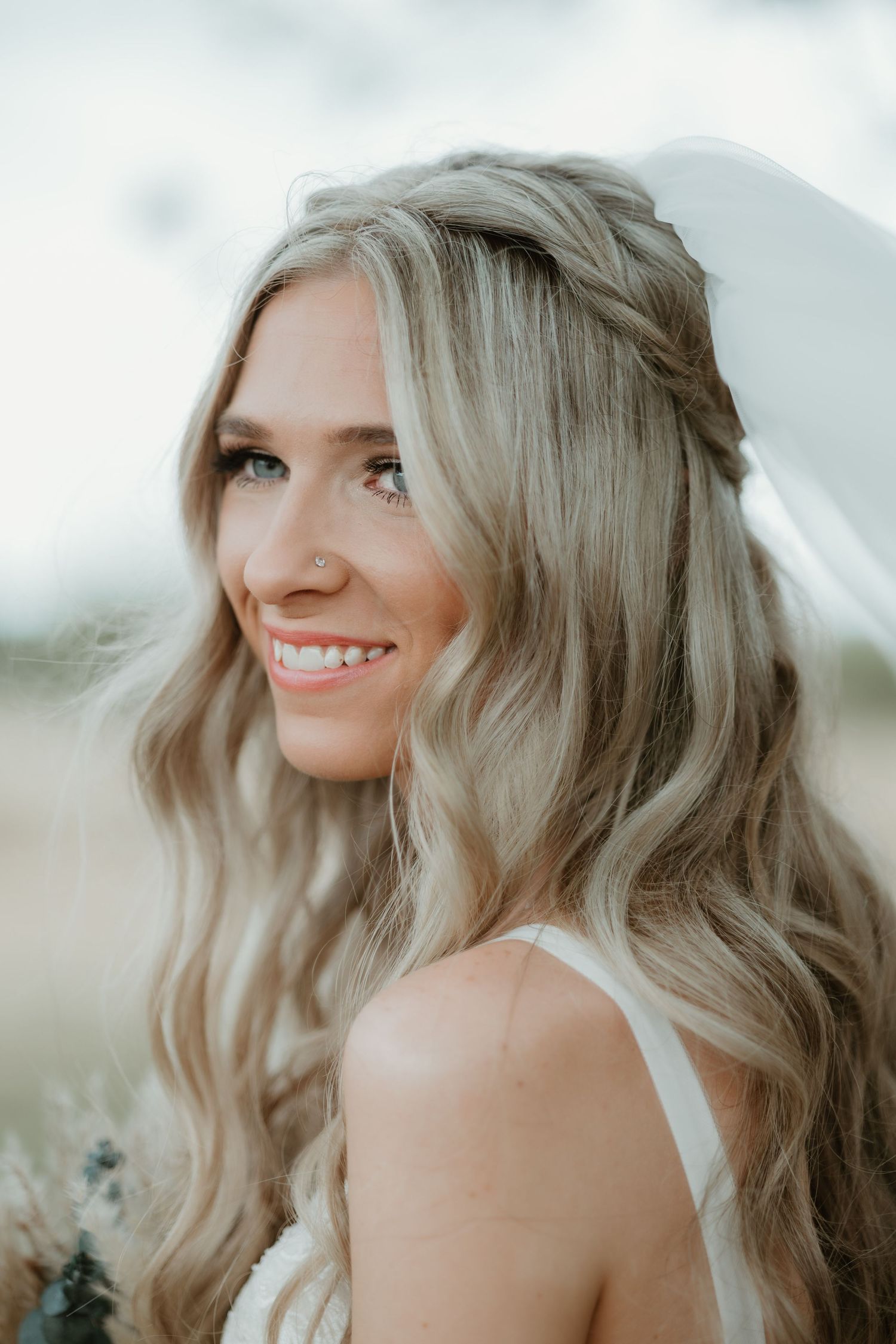 A bride with long wavy blonde hair styled with a delicate braid smiles in an outdoor wedding portrait.