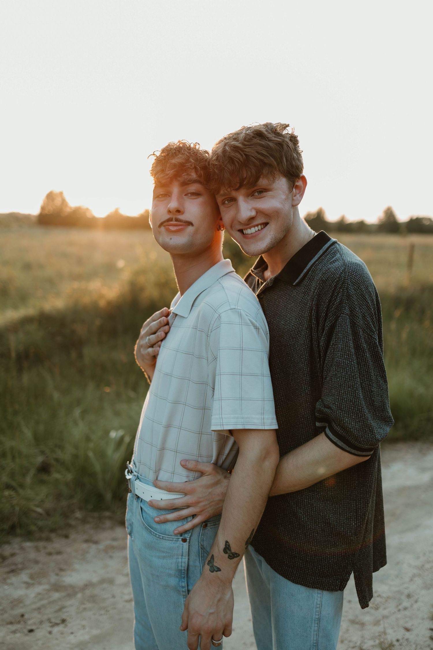 Two people embrace on a dirt path during sunset, with golden light illuminating a grassy field in the background.