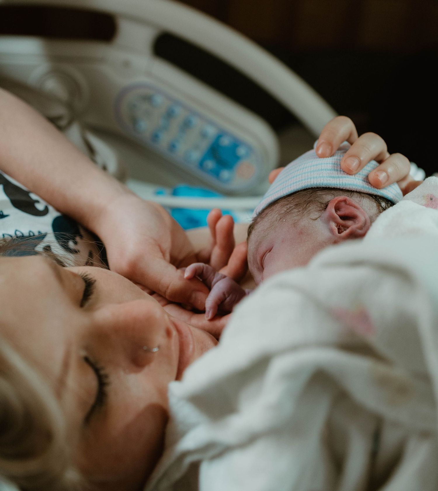 A newborn baby rests peacefully on mother in hospital bed with medical equipment visible in the background.