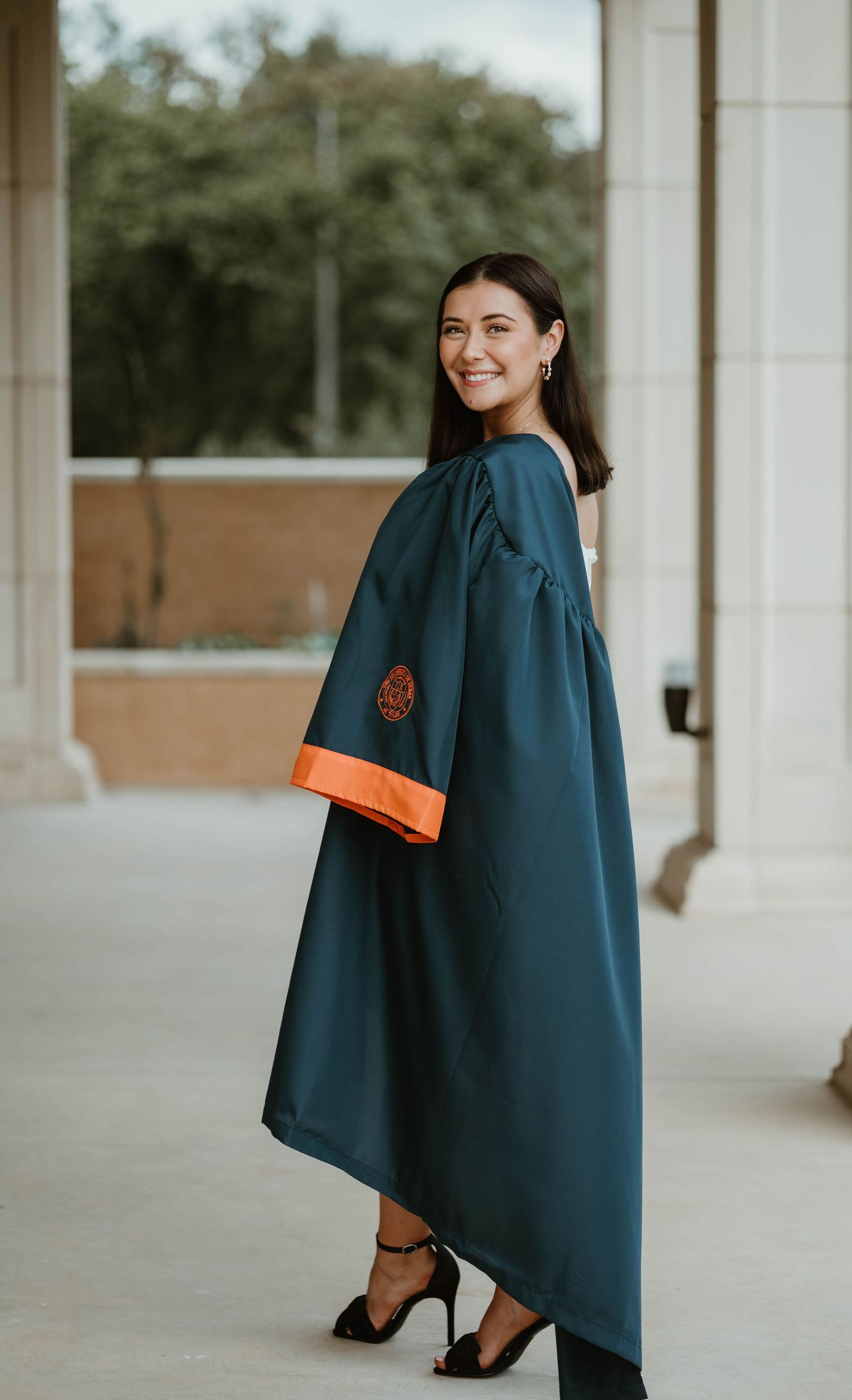 Graduate in teal academic gown poses in elegant black heels showing graduation fashion details.