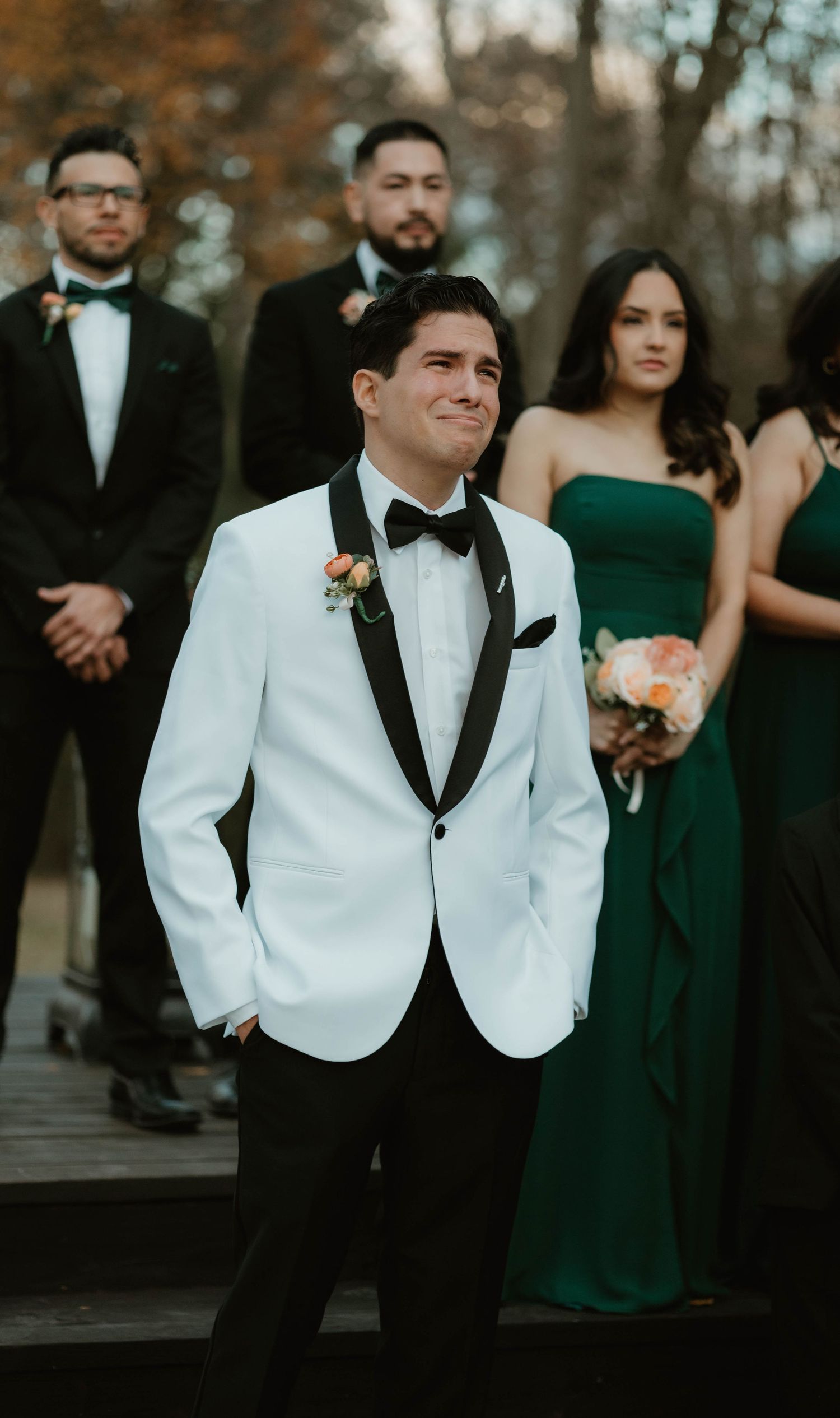 Wedding party in elegant attire with groom in white tuxedo jacket and bridesmaids wearing emerald green gowns outdoors.