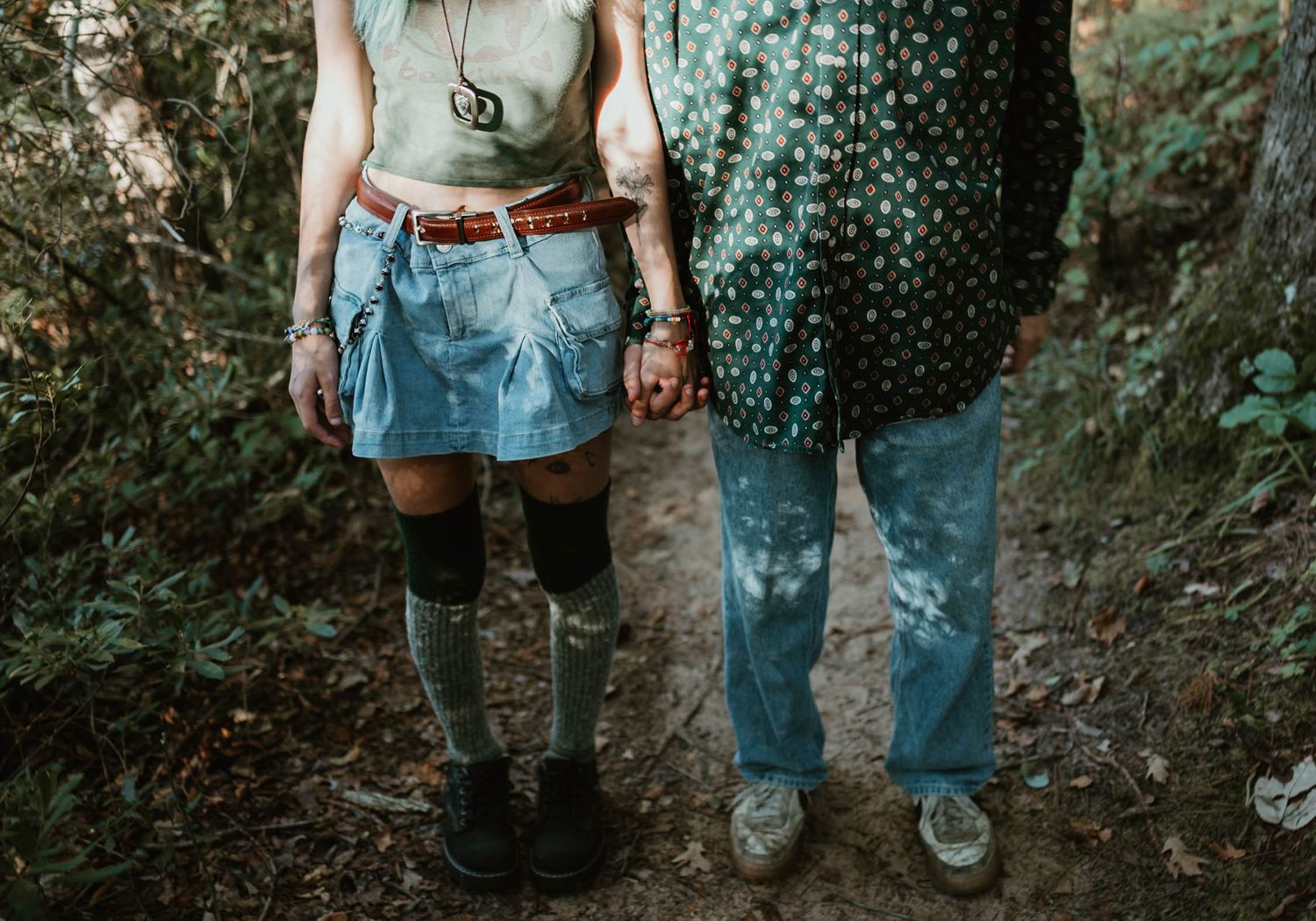 Two people in contrasting denim outfits stand together on a forest path in casual outdoor attire.