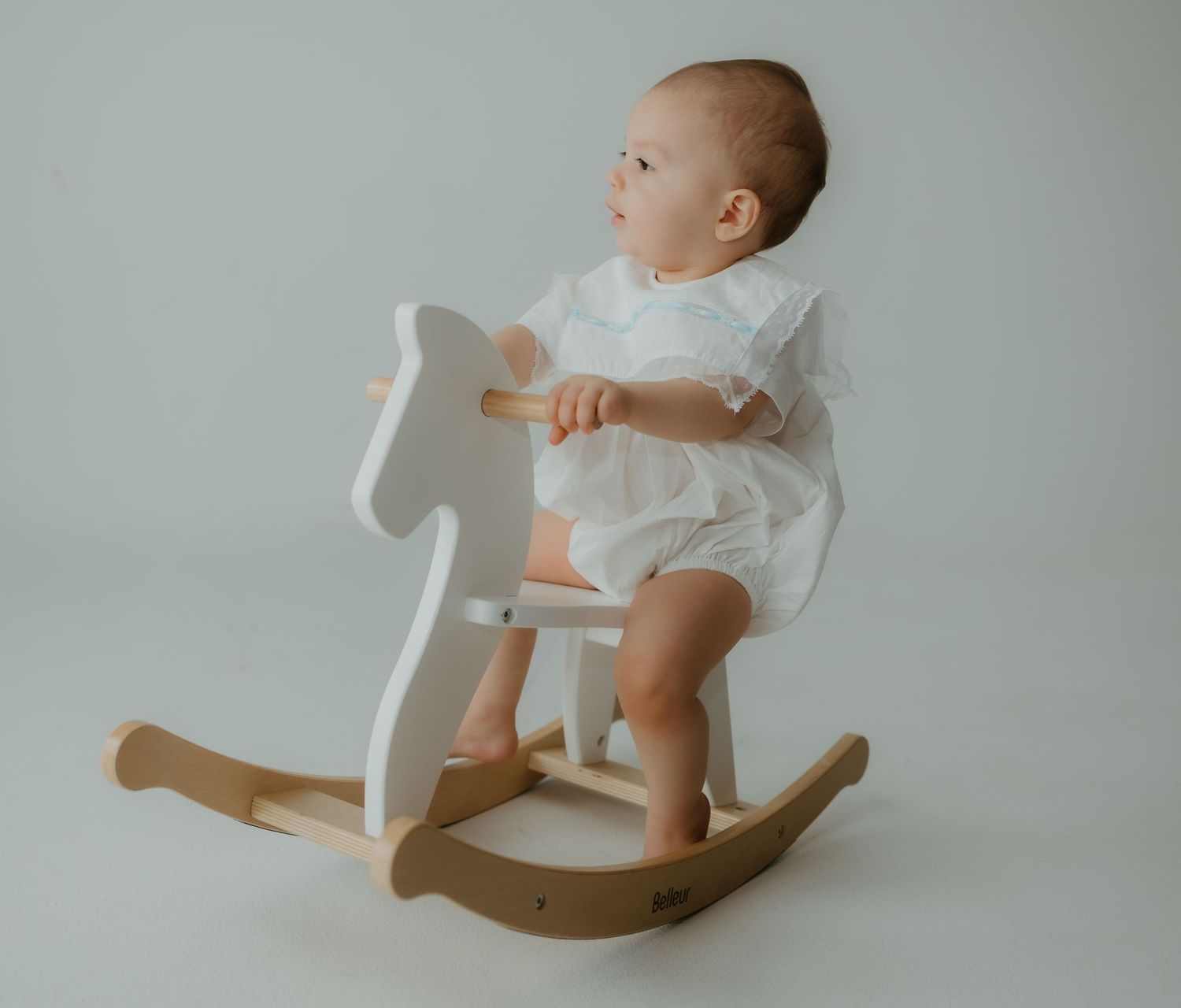 An infant in white clothing plays on a minimalist white wooden rocking horse against a neutral gray backdrop.