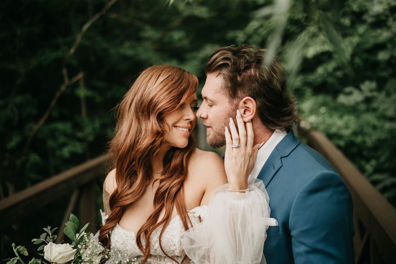 A romantic couple shares an intimate moment while holding white flowers on a wooden bridge surrounded by lush greenery.