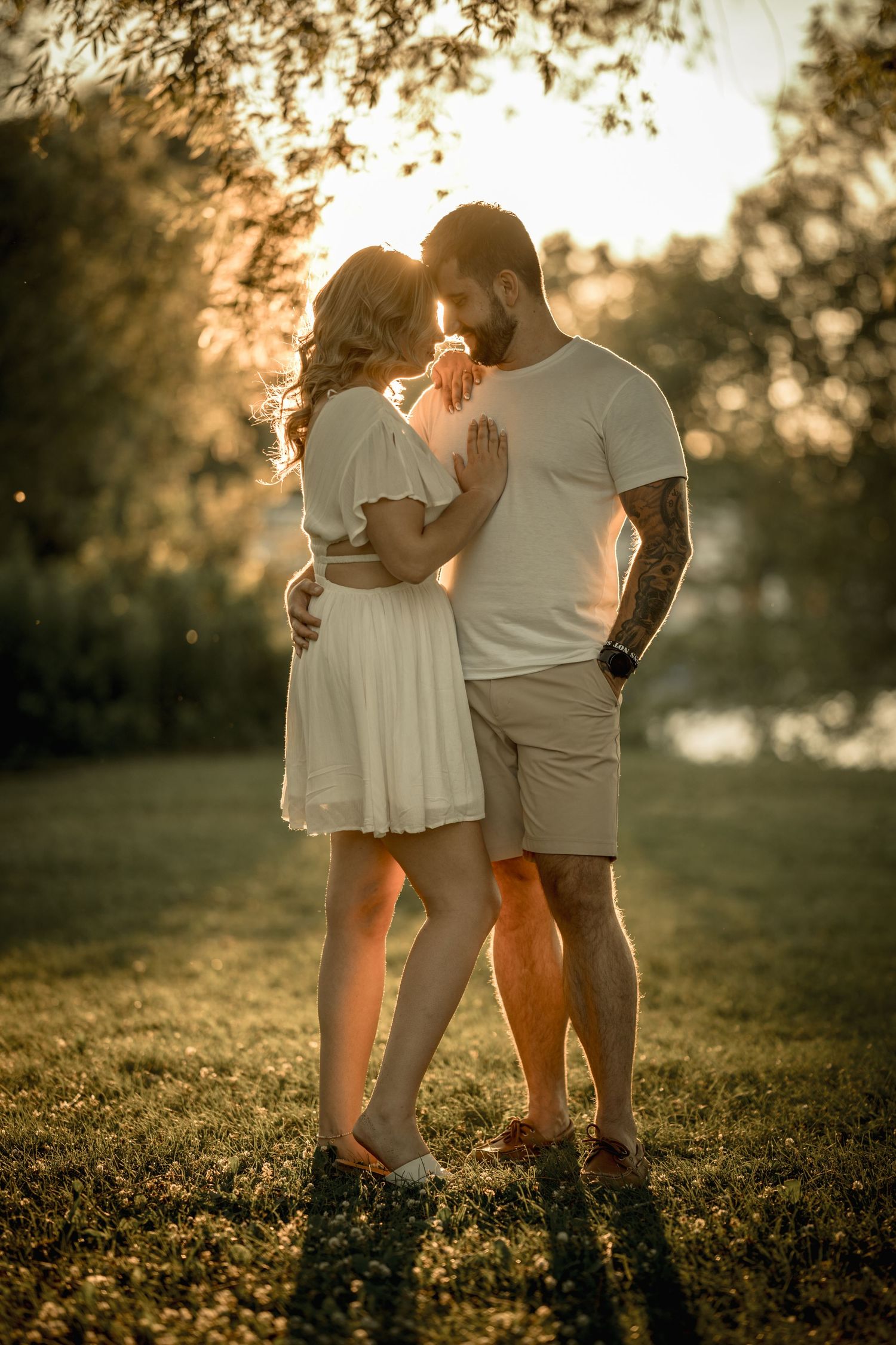 Romantic couple embraces in soft golden sunset light in a grassy park setting.