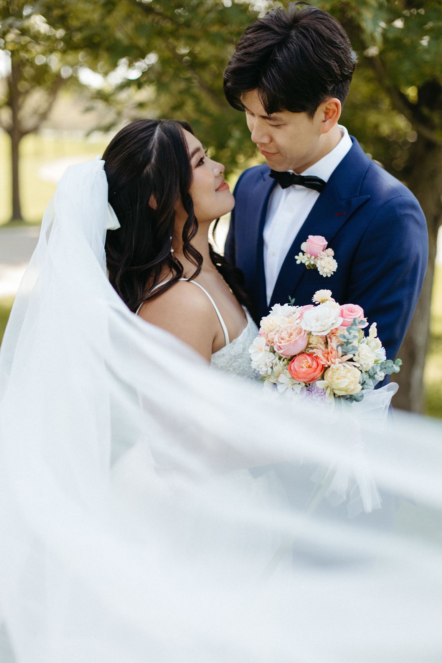 A romantic wedding photo of a couple embracing outdoors, with the bride holding a bouquet of pink and peach flowers.