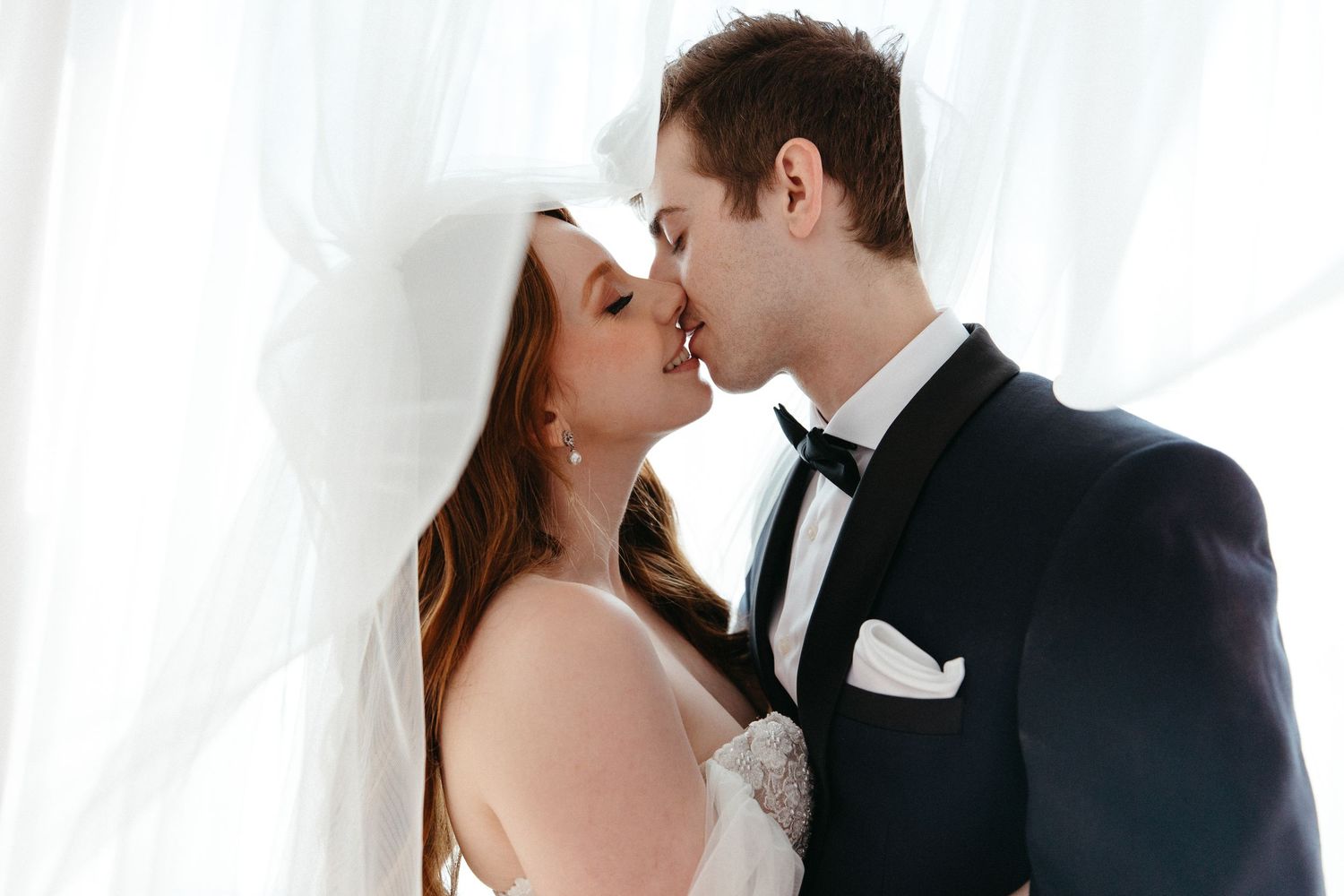 A romantic bride and groom share an intimate kiss against a white curtain backdrop.