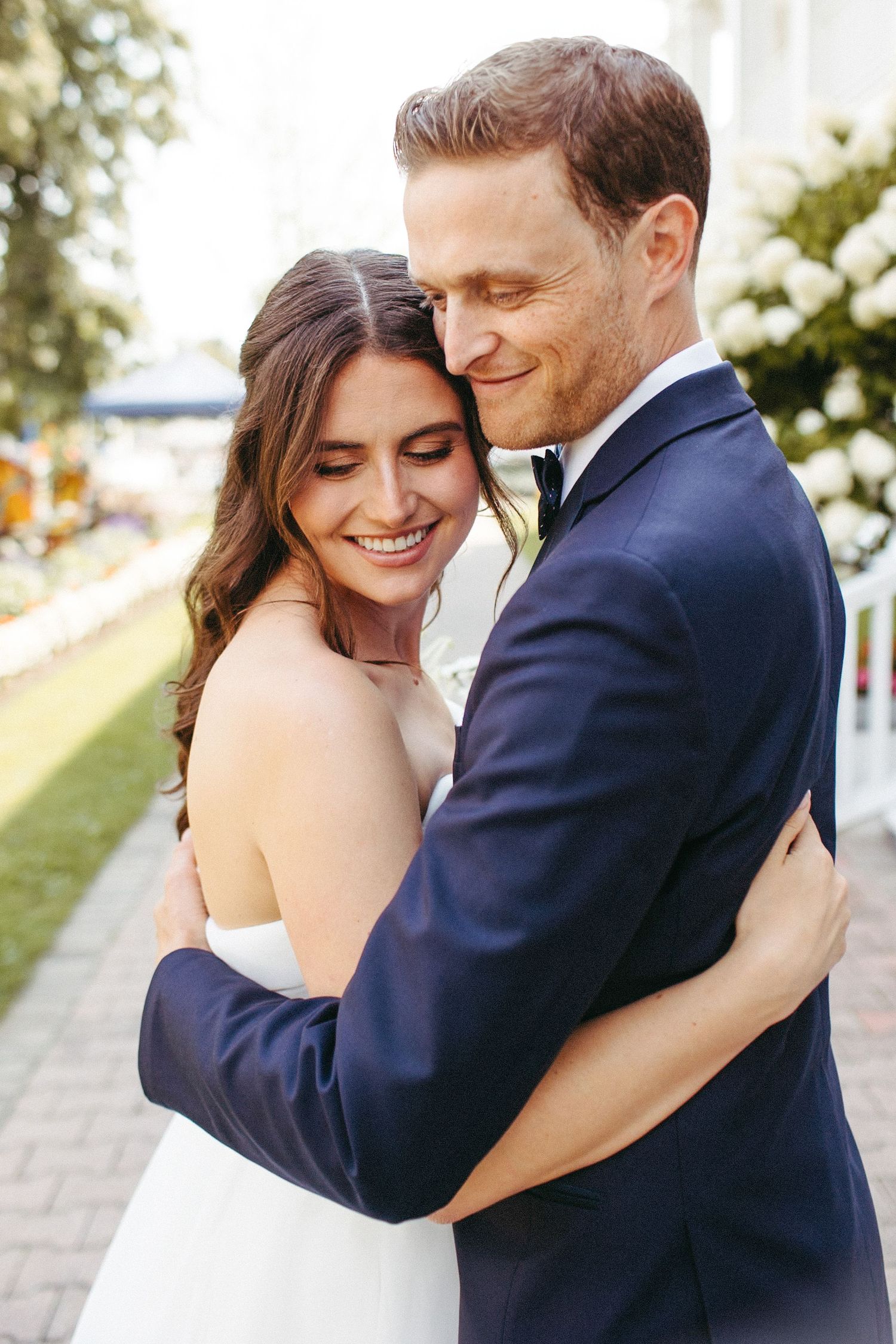 A couple in wedding attire embrace outdoors on a sunny day with greenery in the background.