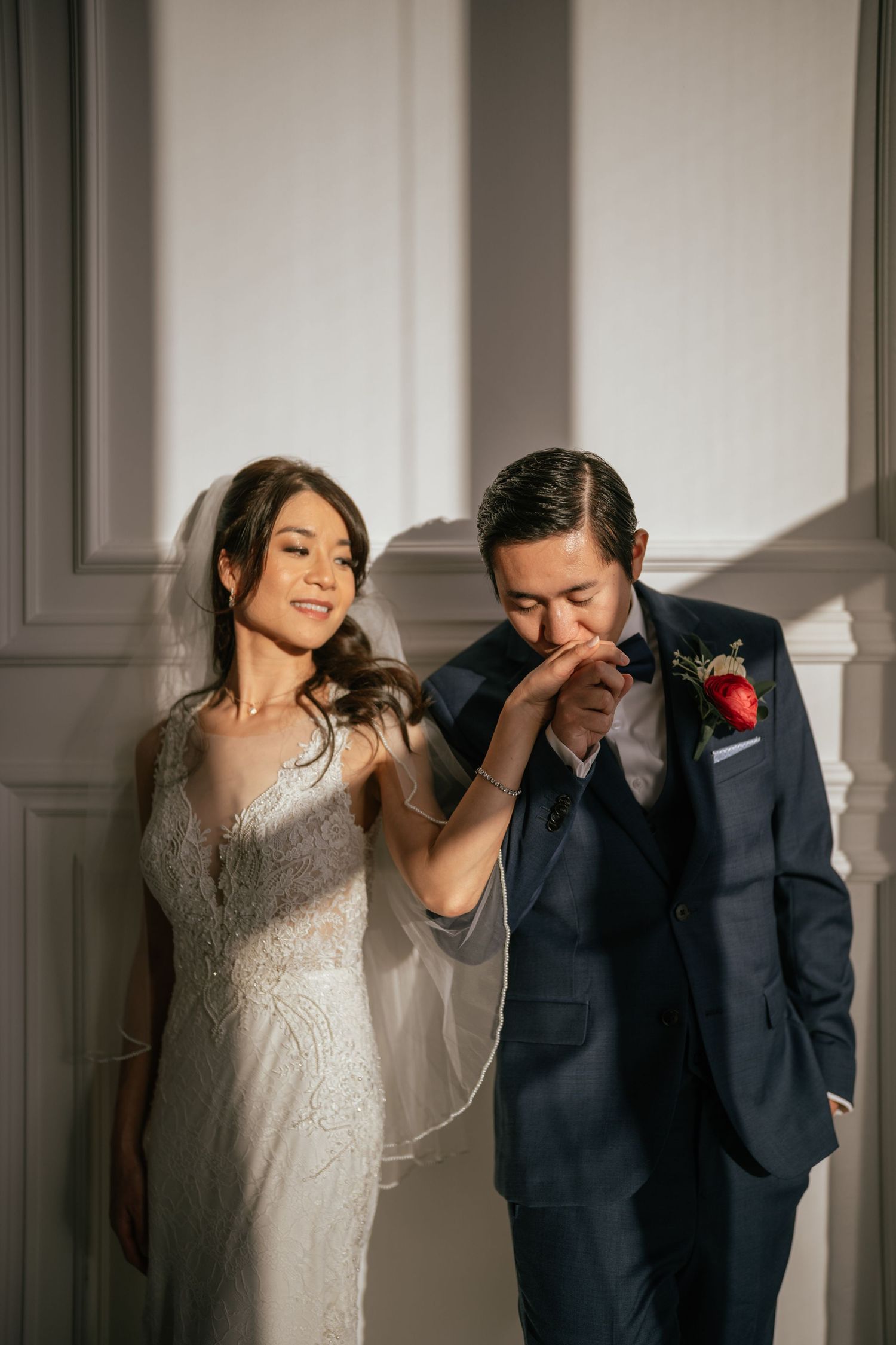 A romantic wedding moment captured in natural light as newlyweds share a tender gesture by a white wall.