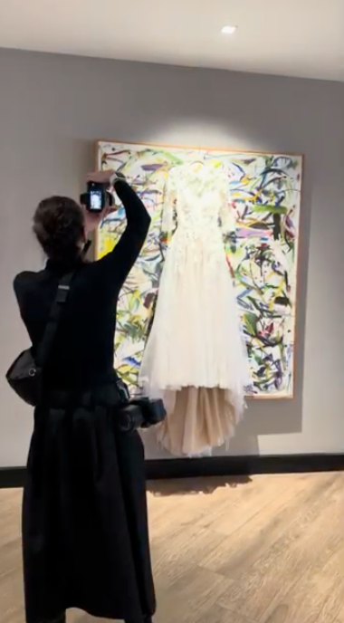 A white wedding dress hangs on display against a colorful abstract art piece as someone takes a photo with their phone.