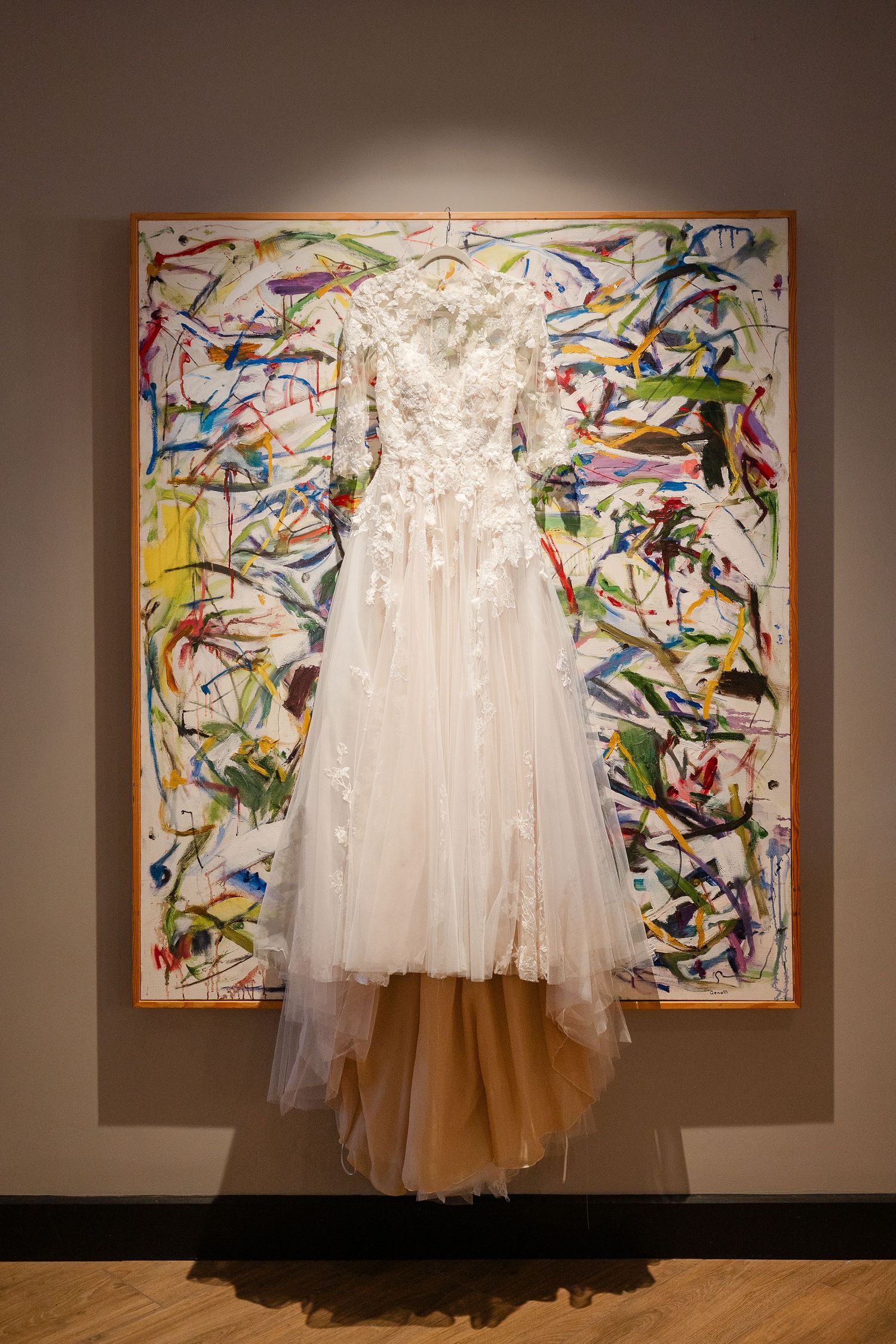 A white wedding dress hangs against a colorful abstract art painting on a gallery wall.
