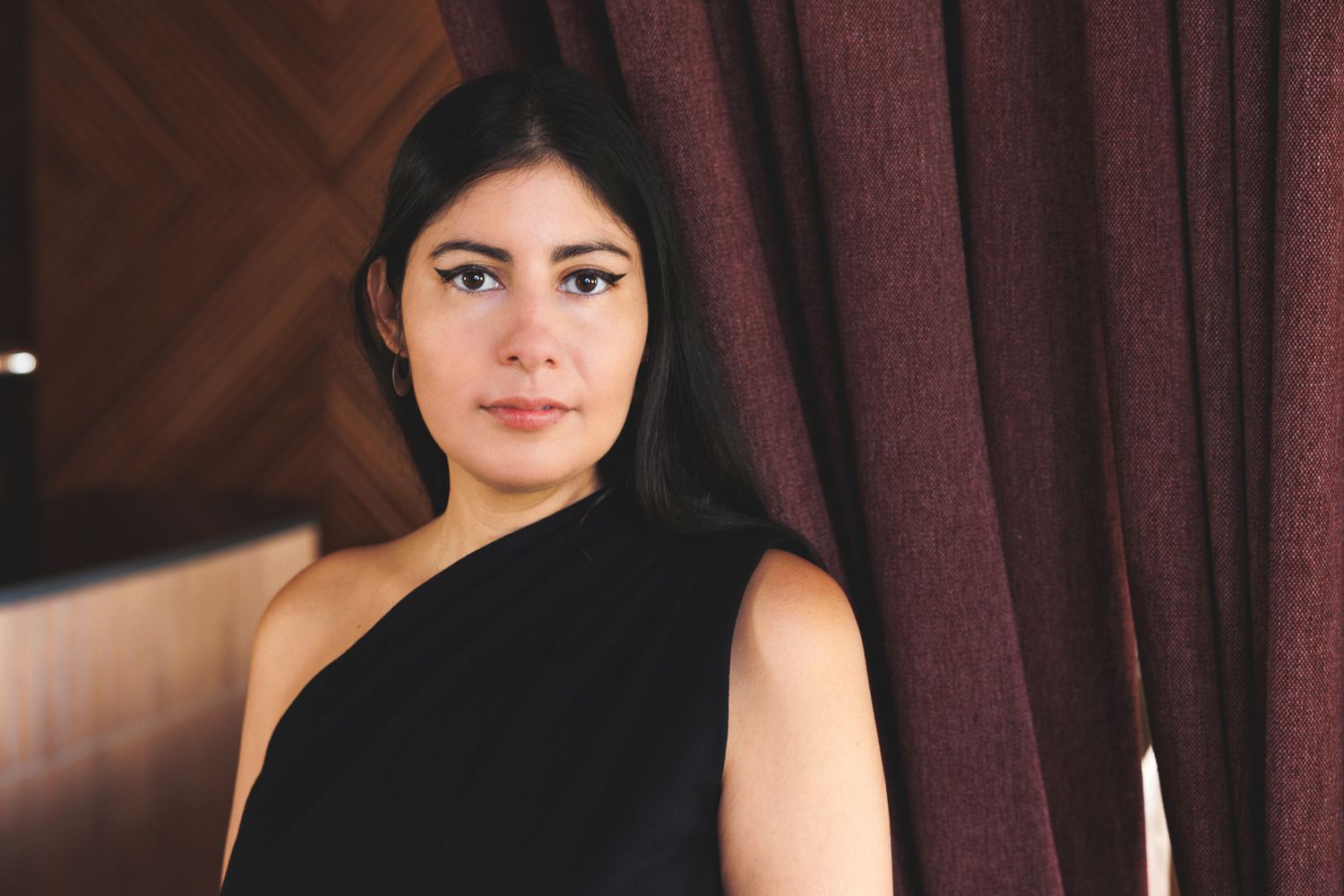 A person poses in a sophisticated one-shoulder black dress against a burgundy curtain backdrop.