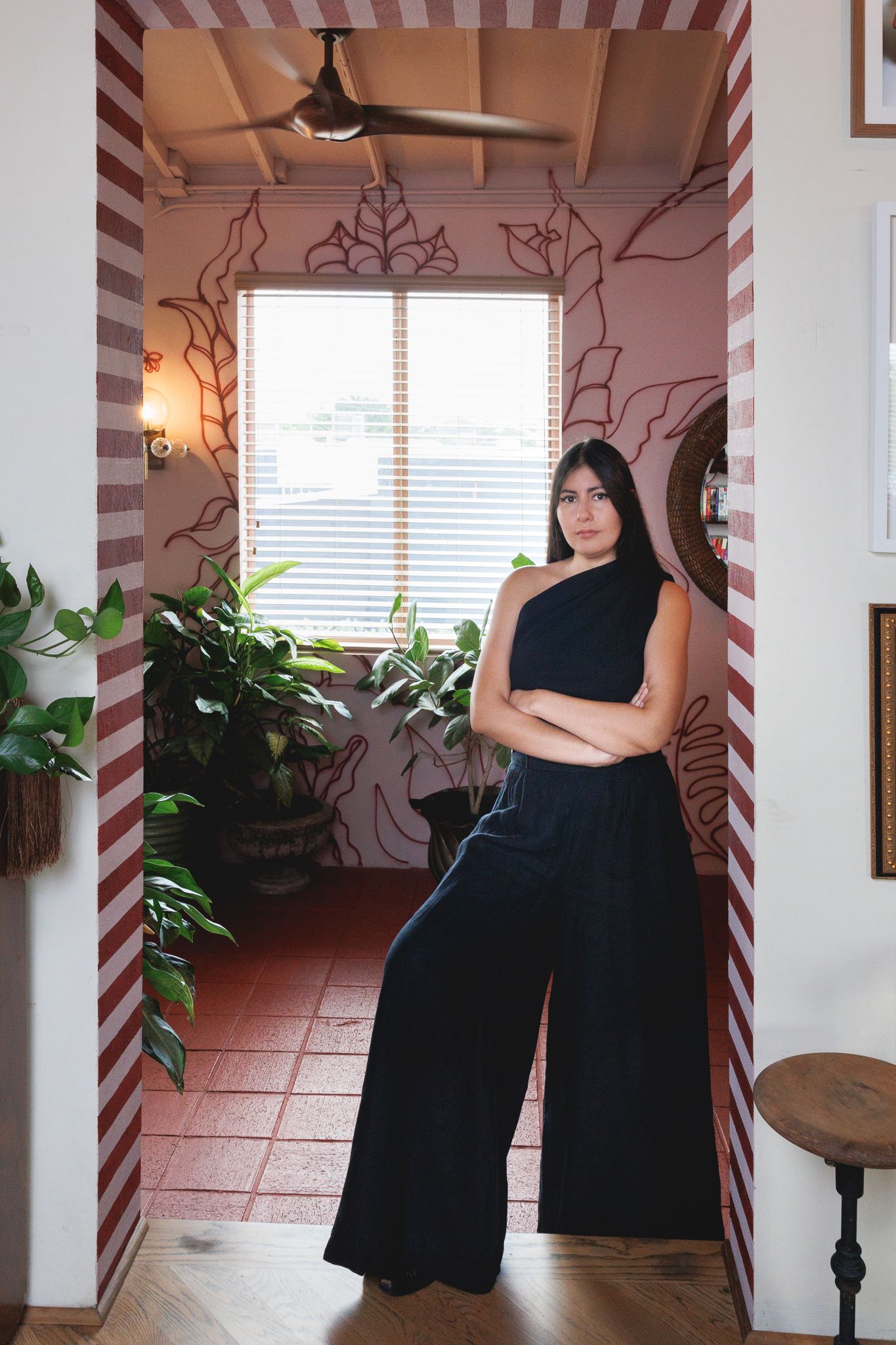 A one-shouldered black jumpsuit is styled in a room with terra cotta floors and decorative wall vines.