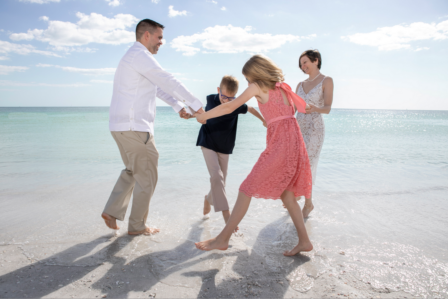 A family hold hands while dancing together on a white sandy beach with blue ocean in background in a miami lifestyle family photos shoot